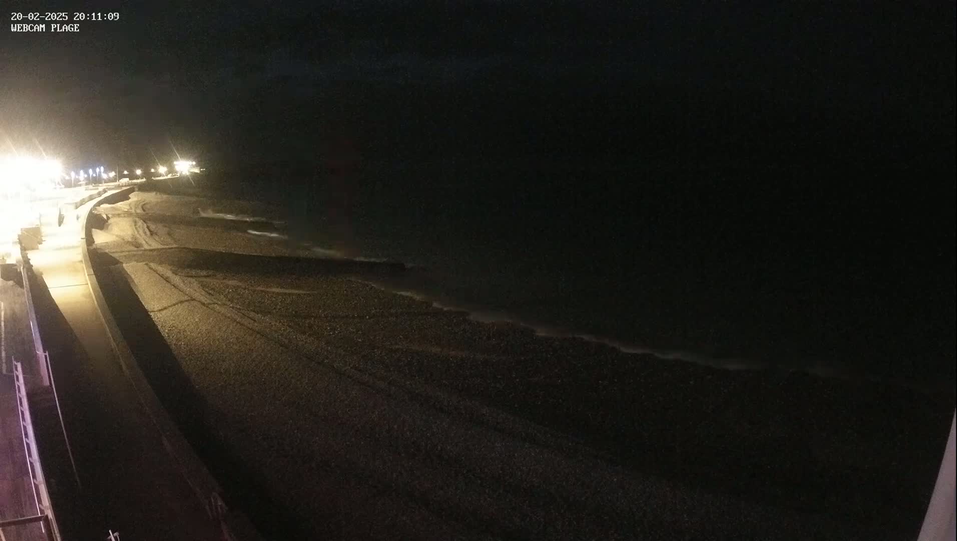 A dark, nighttime view of a pebble beach and a paved walkway, illuminated by streetlights, with gentle waves lapping at the shore.