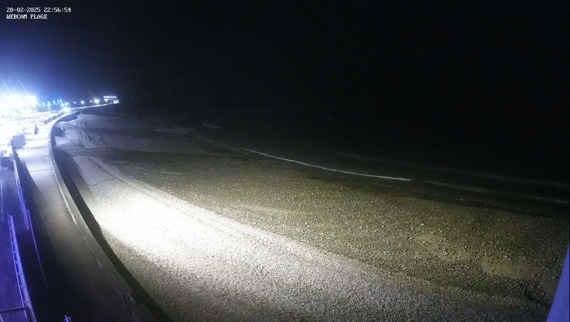 A nighttime view of a pebble beach and a paved walkway, illuminated by artificial lights, with calm waves gently lapping the shore.