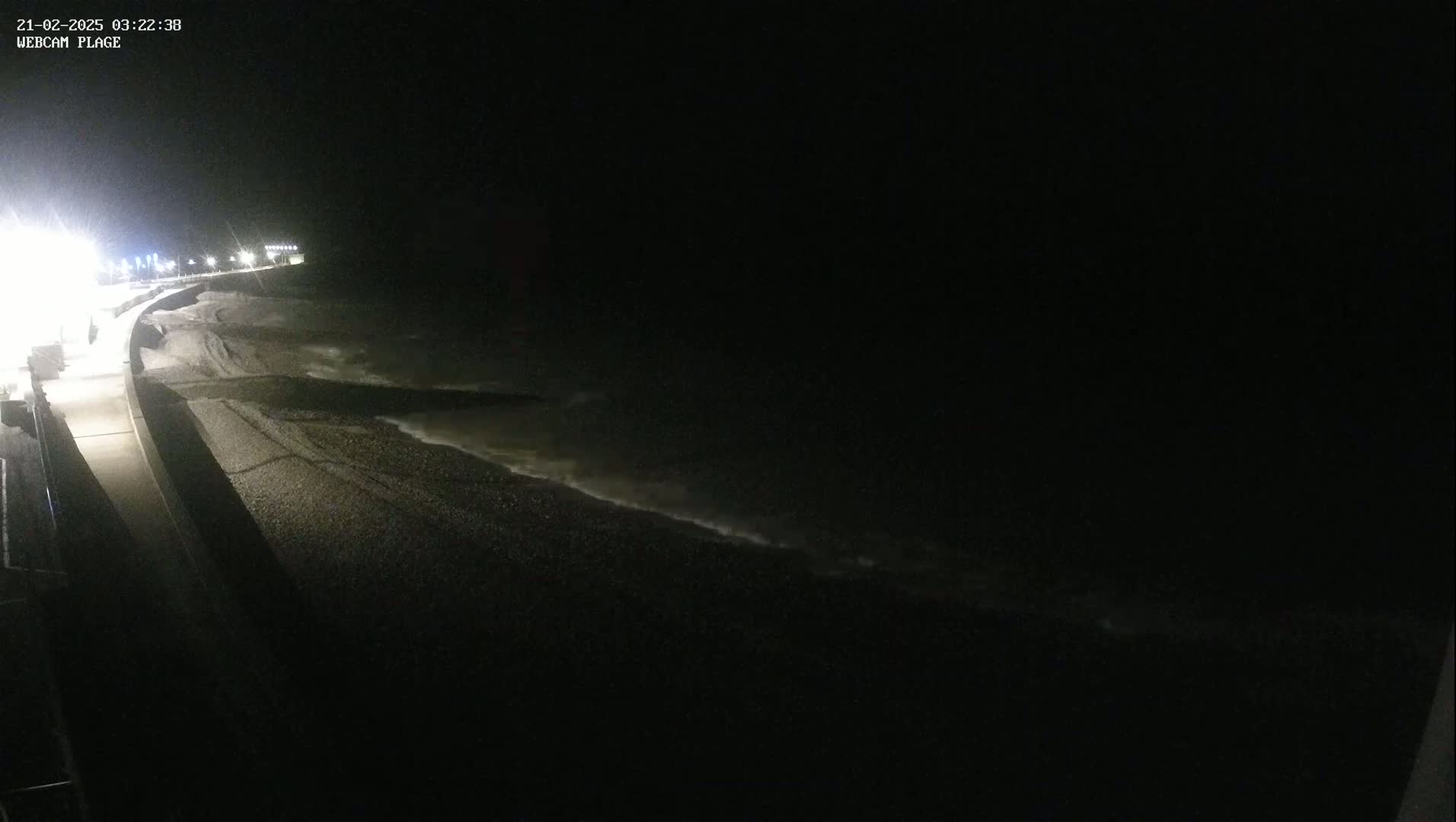 A dark, nighttime view of a pebble beach with gently lapping waves under a clear, black sky.