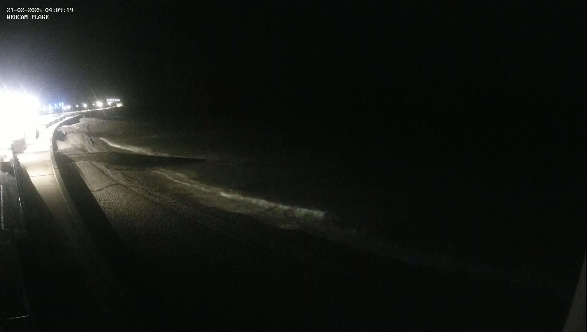 A dark, nighttime view of a pebble beach and ocean with small waves lapping the shore, illuminated by lights along a nearby walkway.