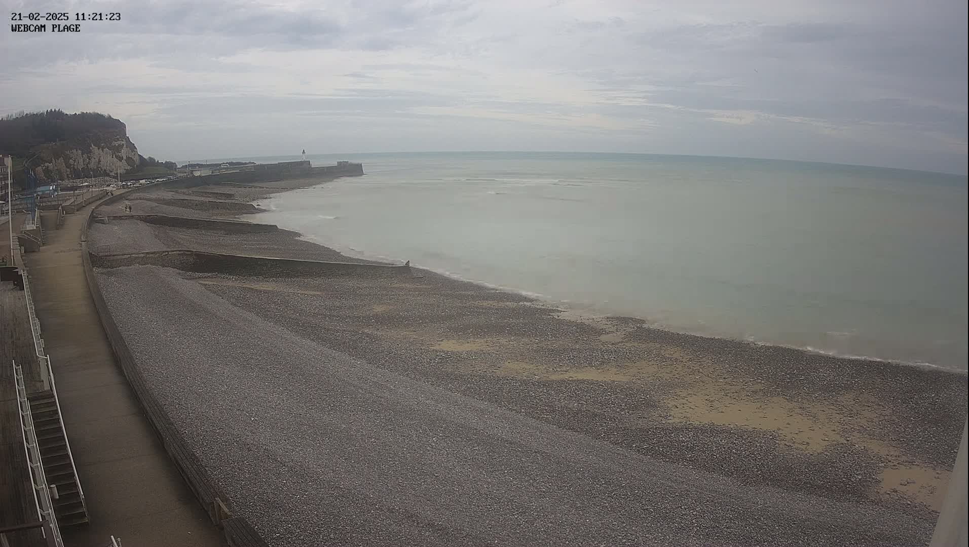 A pebble beach stretches along a calm, overcast sea, next to a paved walkway and a cliff face.