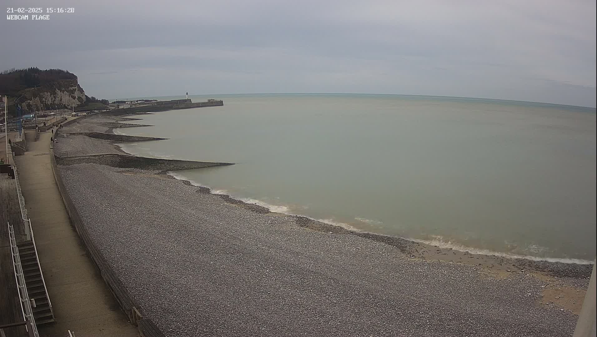 A pebble beach stretches alongside a paved walkway, leading to a low sea wall and a calm, grey sea under an overcast sky.