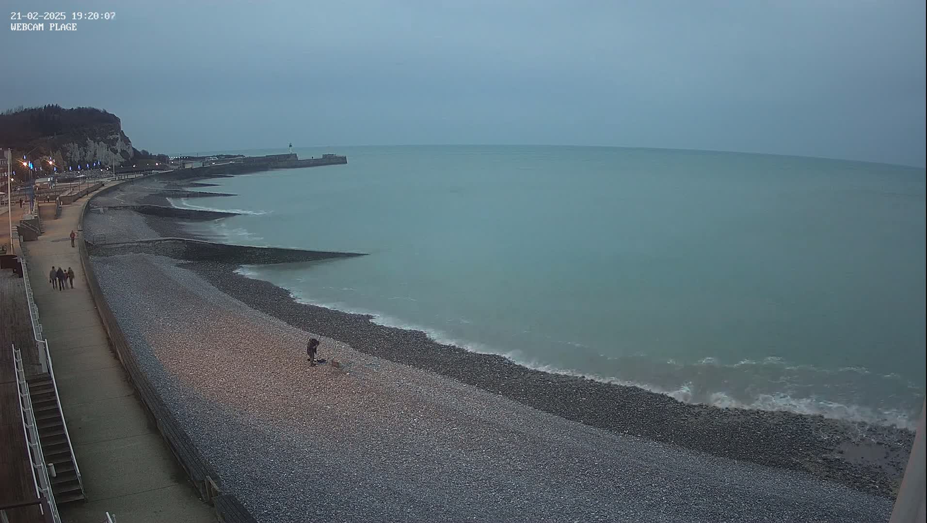 A pebble beach stretches along a calm, gray-green sea under an overcast sky, with a few people walking along a promenade and one person near the water's edge.
