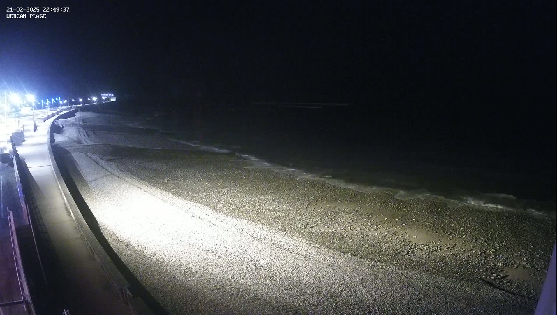 A nighttime, high-angle view shows a pebble beach with gentle waves lapping the shore, next to a paved walkway lit by streetlights.