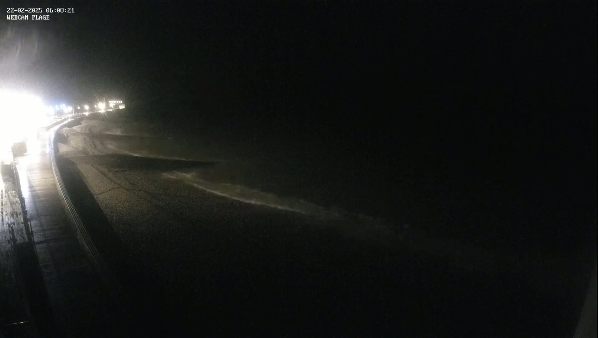 At night, waves crash on a dark, pebble beach next to a rain-slicked road.