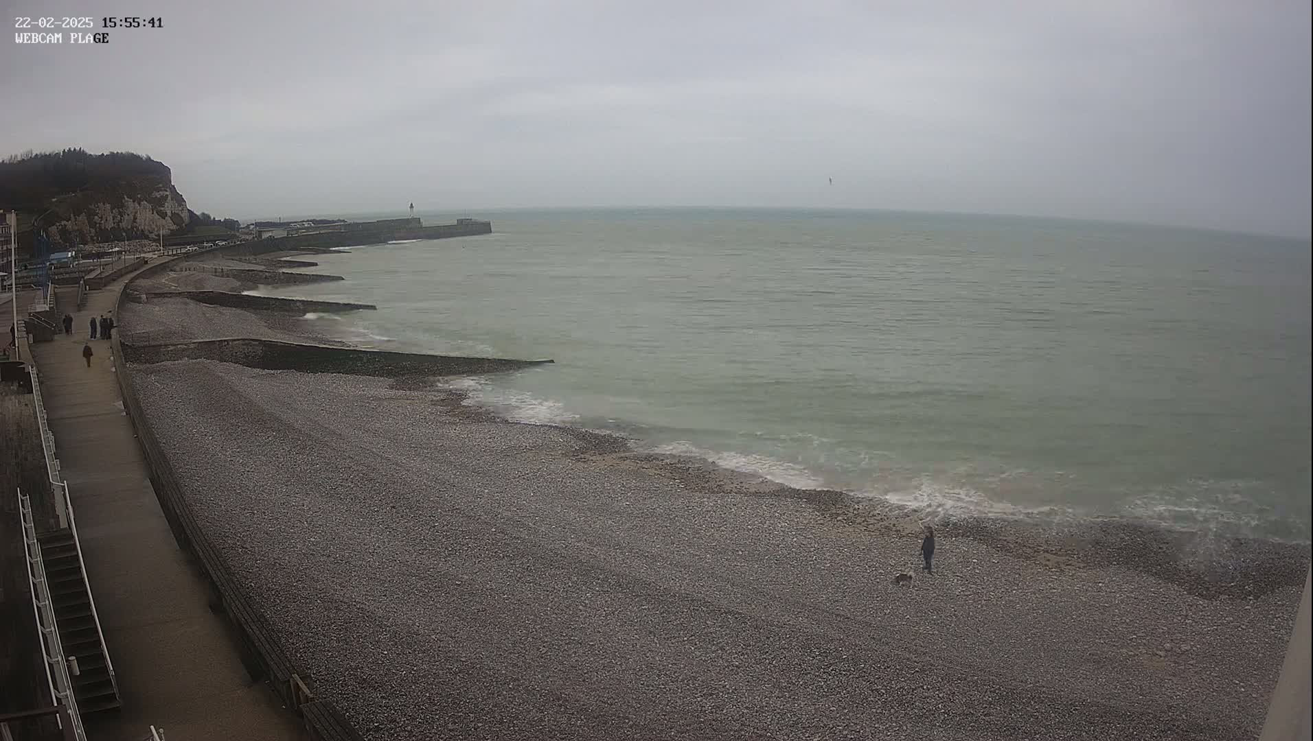 A pebble beach stretches along a calm, gray sea under an overcast sky, with a paved walkway and white cliffs visible in the background.