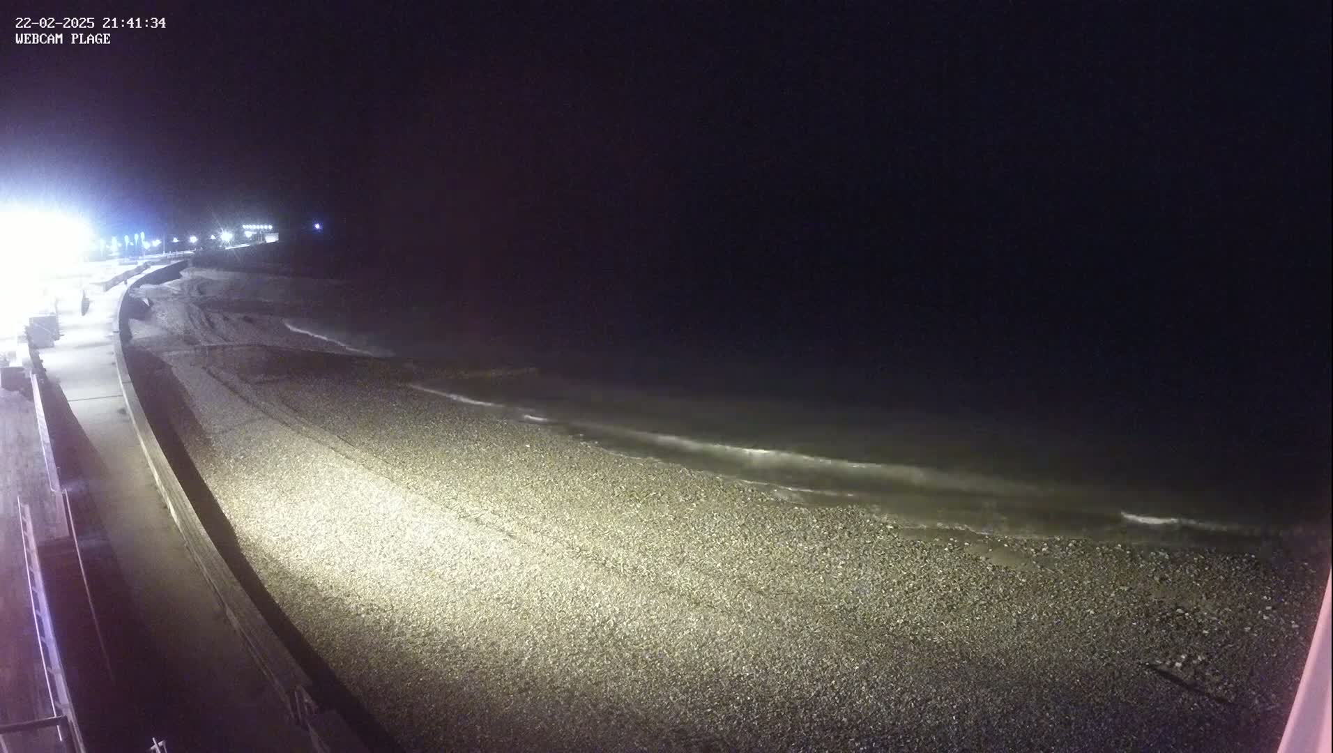 A dark, pebble beach is gently lapped by waves at night, illuminated by artificial lights along a nearby walkway.