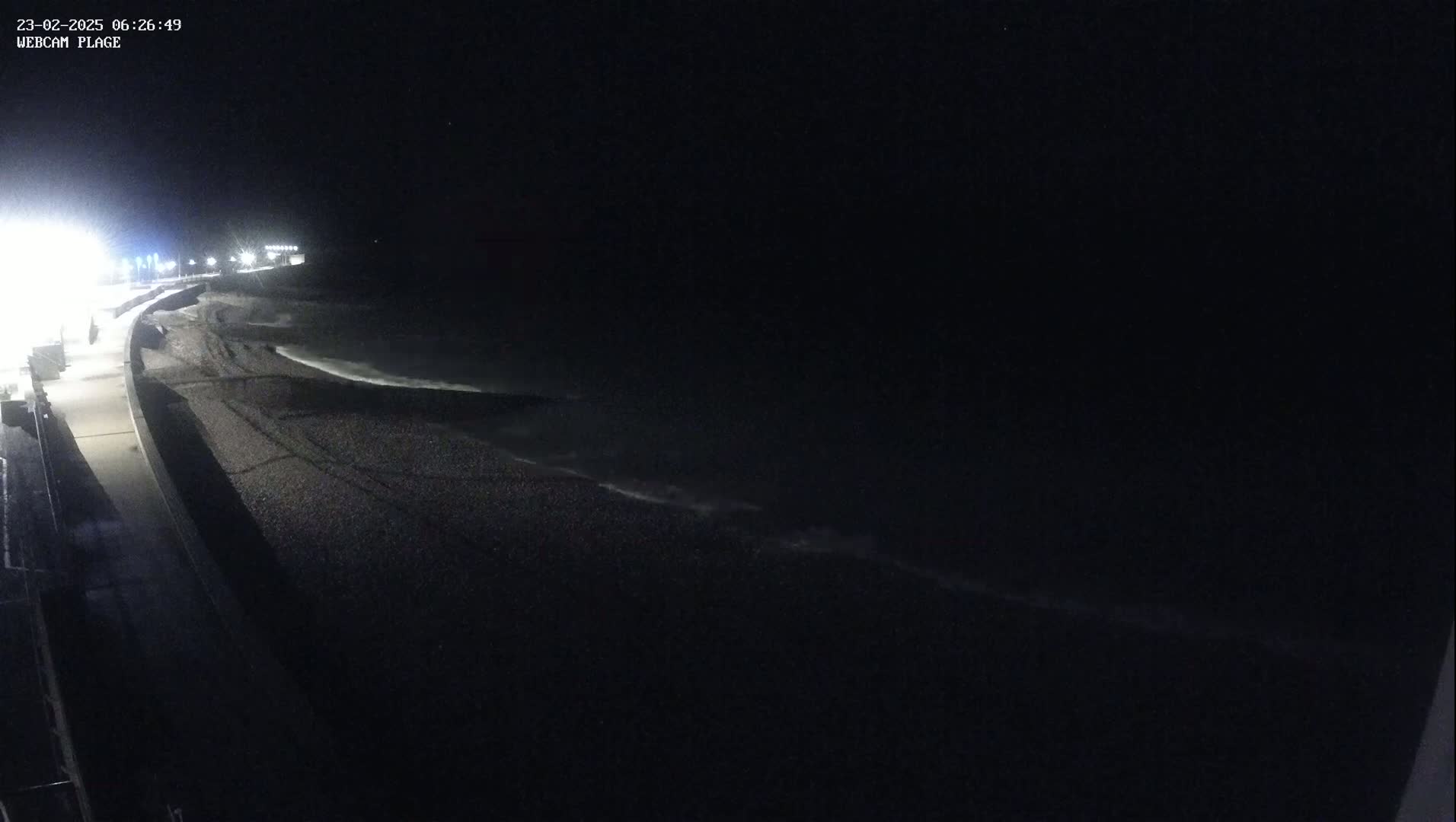 A dark, nighttime view of a pebble beach and ocean with gently lapping waves under a clear, starless sky.