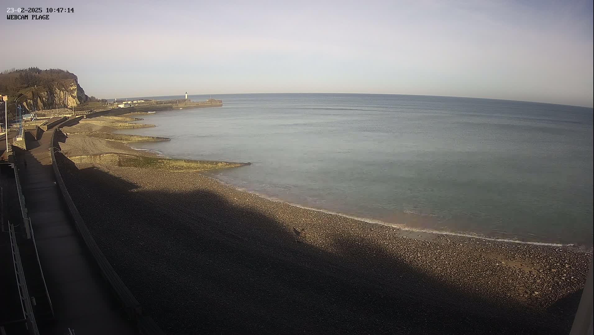 A pebble beach stretches out to a calm, gray-green sea under a pale, clear sky, with a cliff and promenade visible in the background.