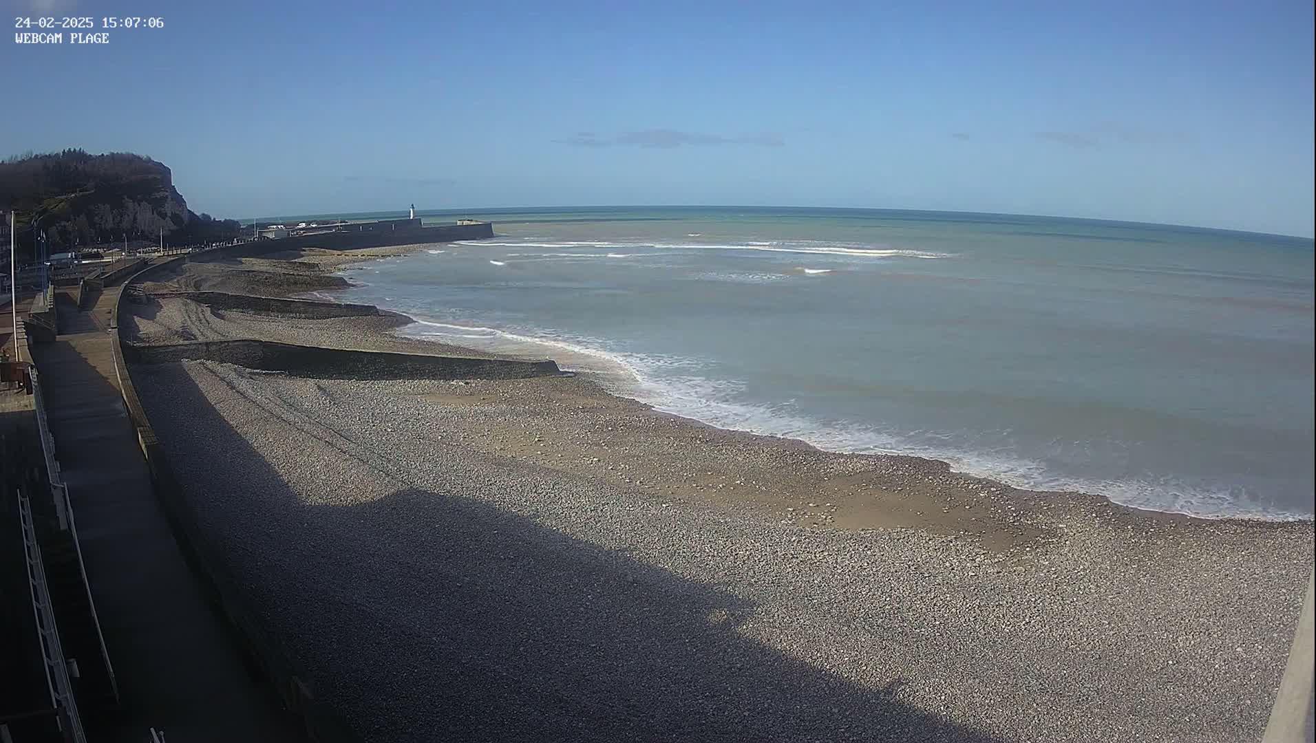 A pebble beach stretches along a calm sea under a clear blue sky, with a walkway and cliff visible in the background.