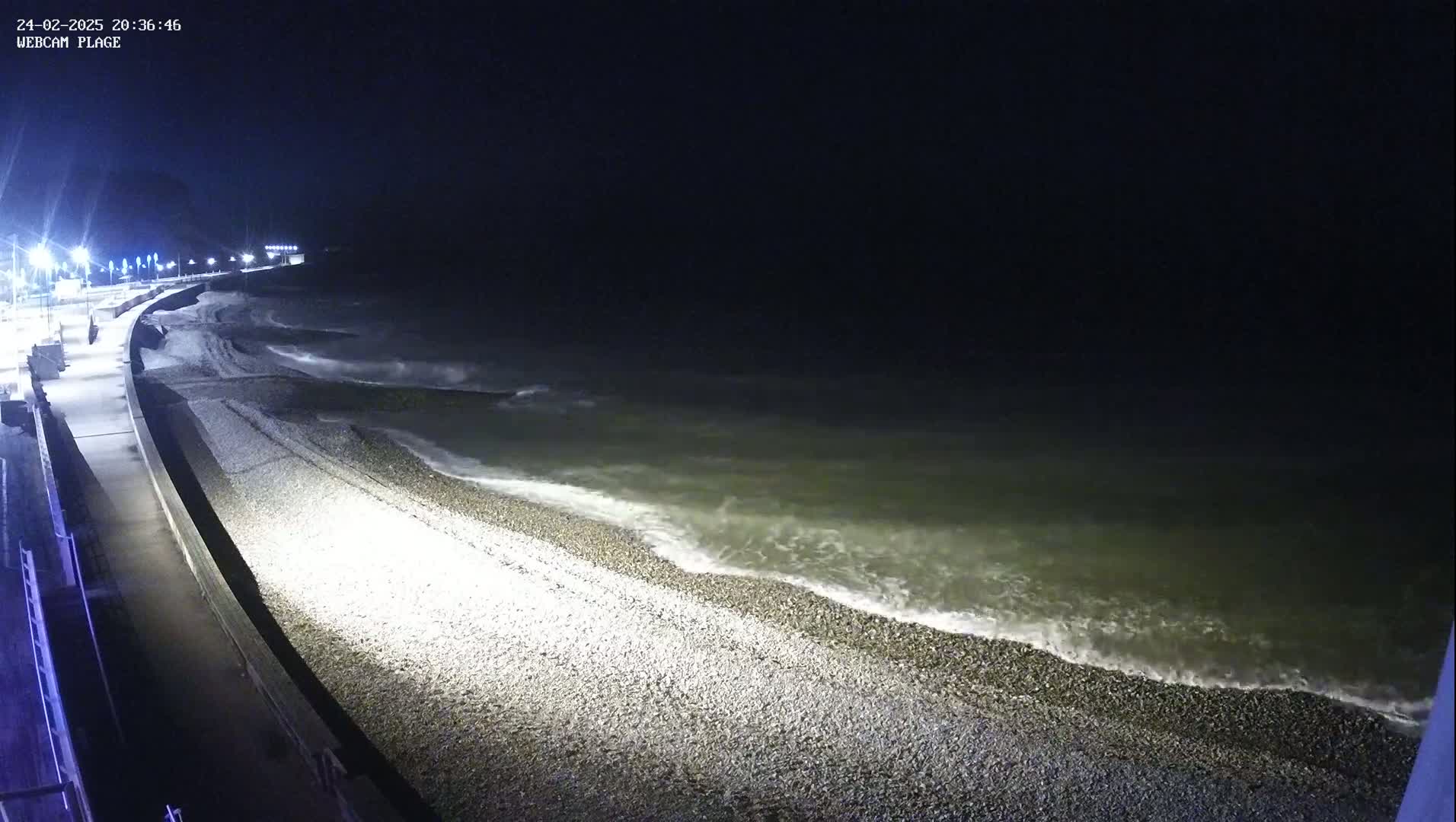 A dark, nighttime beach scene shows gently lapping waves on a pebble shore, illuminated by artificial lights along a paved walkway.
