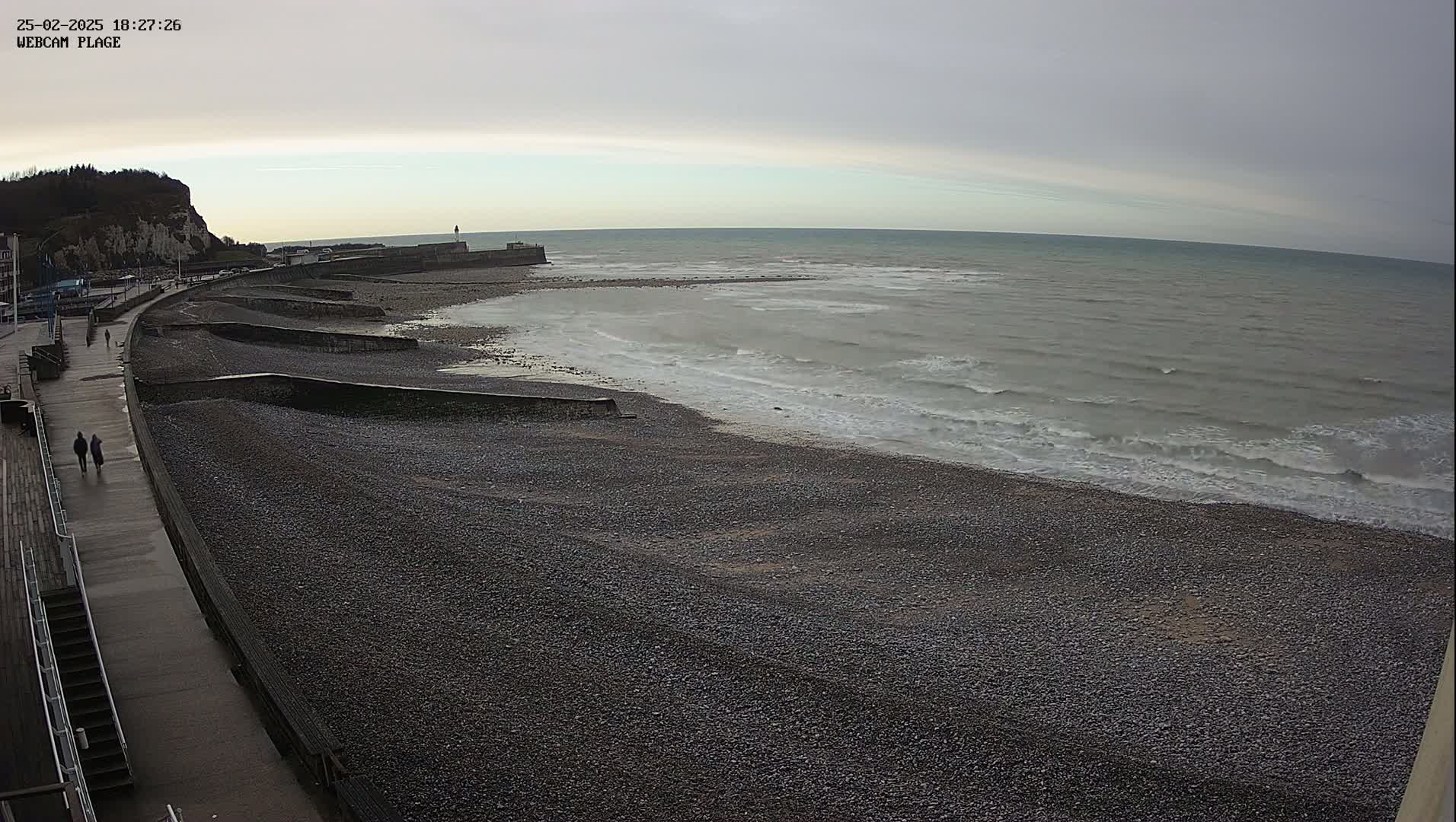 A pebble beach stretches alongside a paved walkway, with gentle waves lapping the shore under an overcast sky.