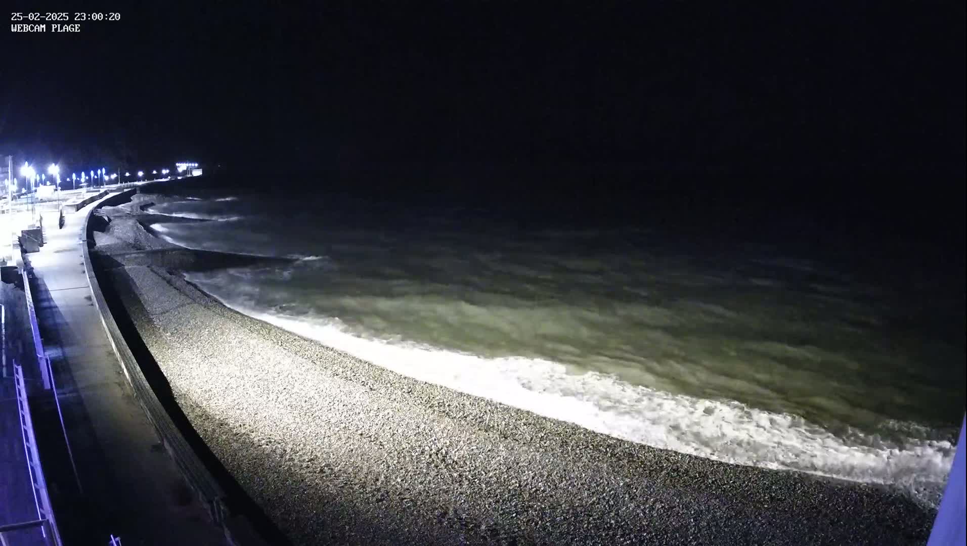 At night, moderately sized waves crash on a pebble beach next to a paved walkway lined with lights.