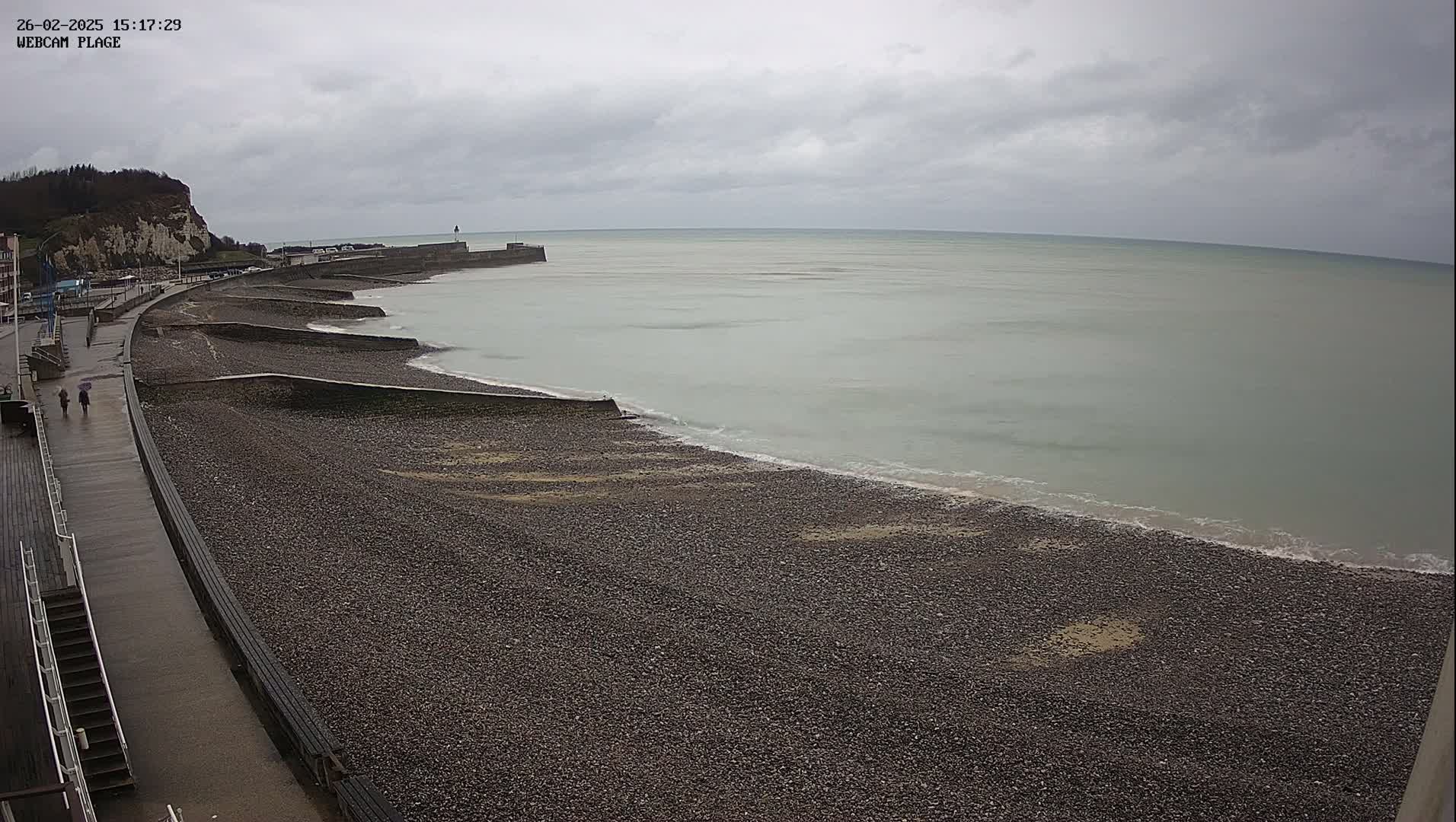 A pebble beach stretches along a calm, gray sea under an overcast sky, with a paved walkway and a cliff visible in the background.