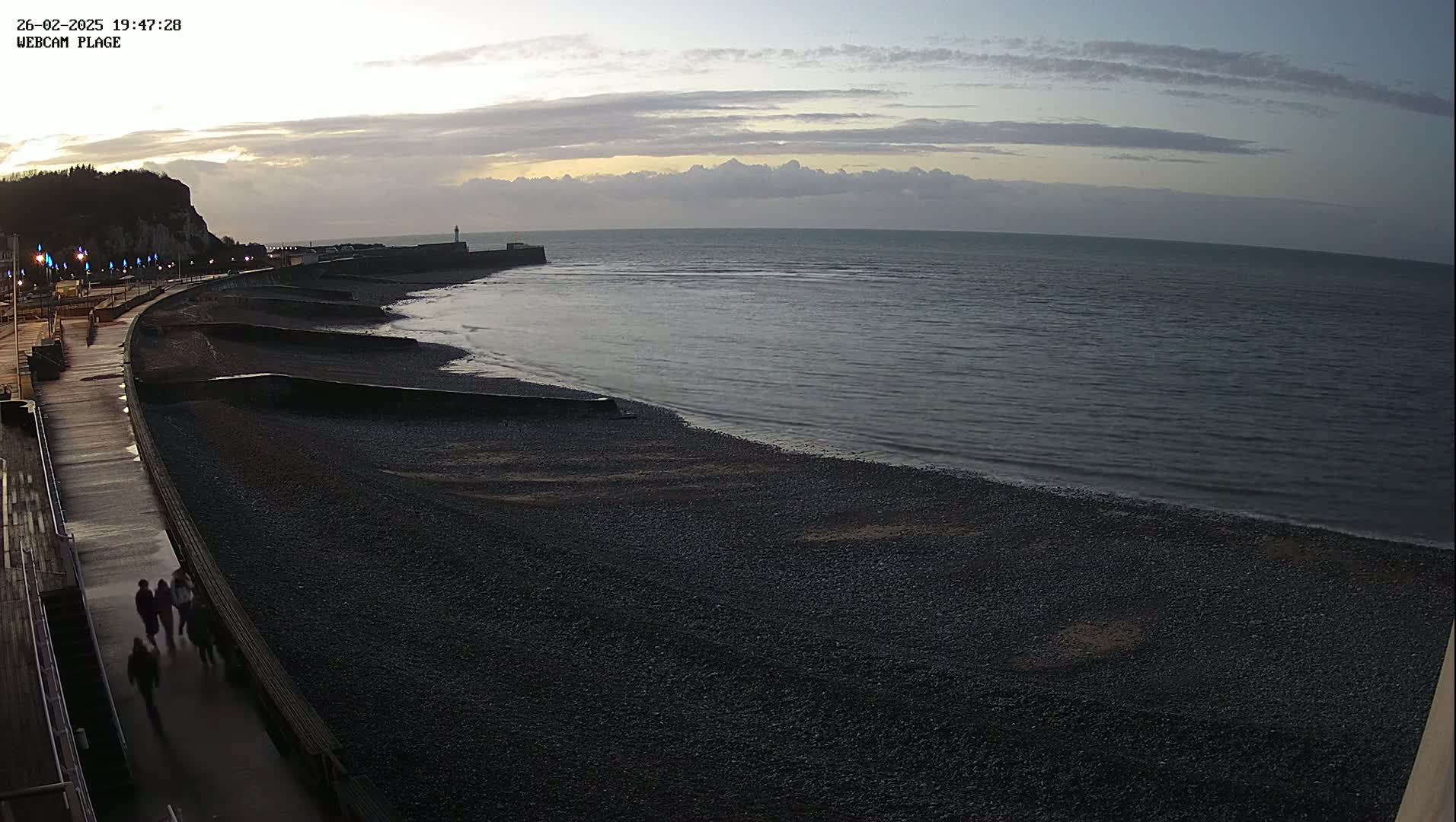 A dark, pebble beach stretches along a curved walkway under a twilight sky with a calm sea in the background.