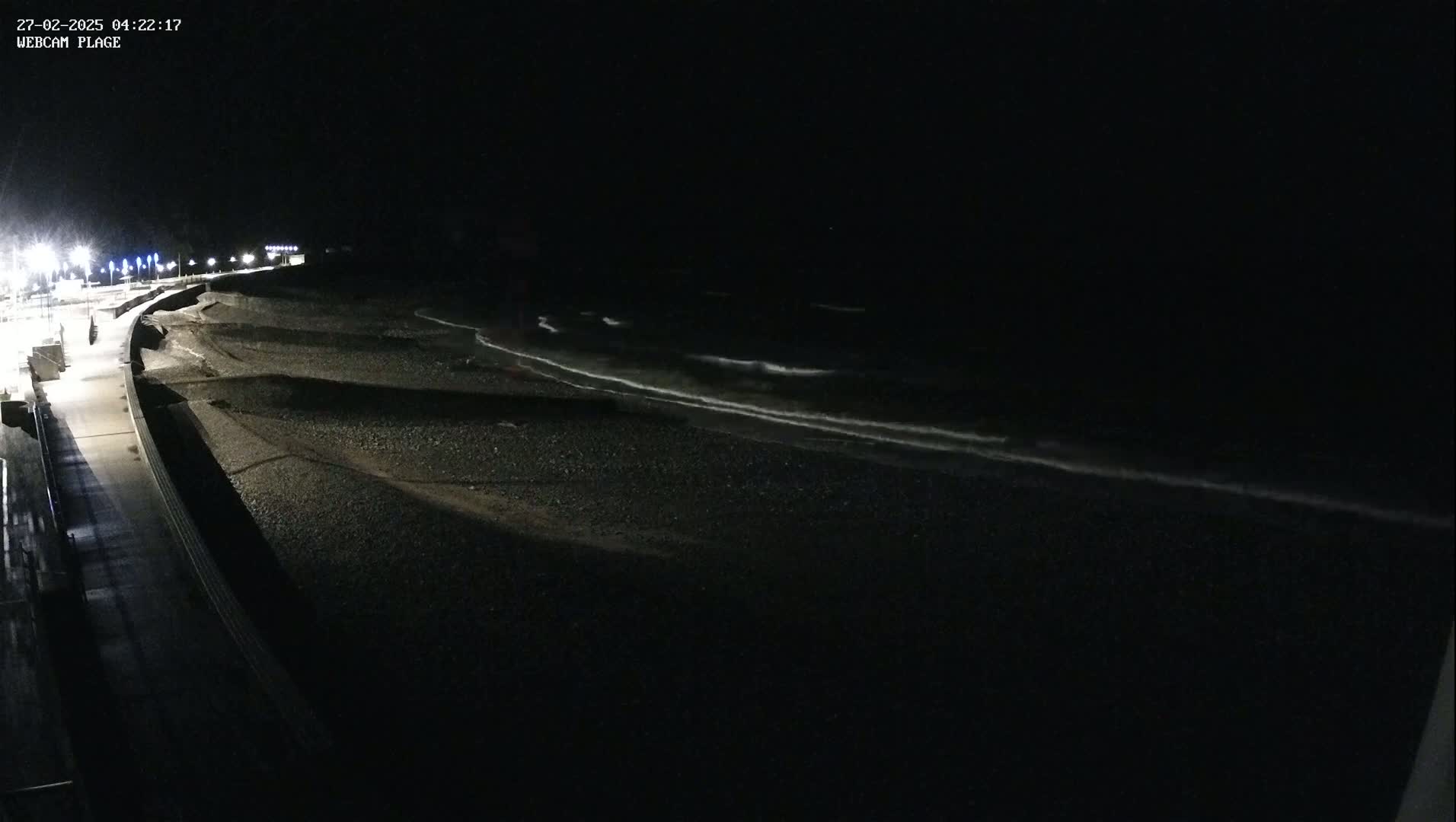 A dark, nighttime view of a pebble beach with gentle waves lapping the shore, illuminated by streetlights along a nearby walkway.