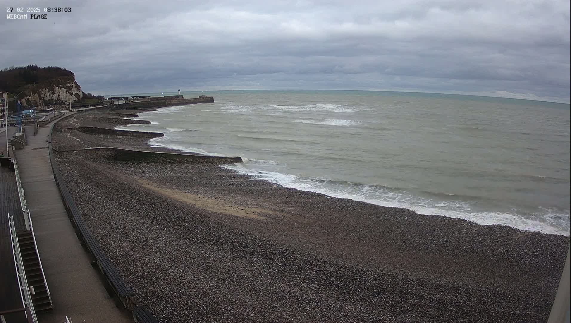 A pebble beach with a walkway and sea wall stretches along a coastline under a cloudy sky, with small waves lapping the shore.