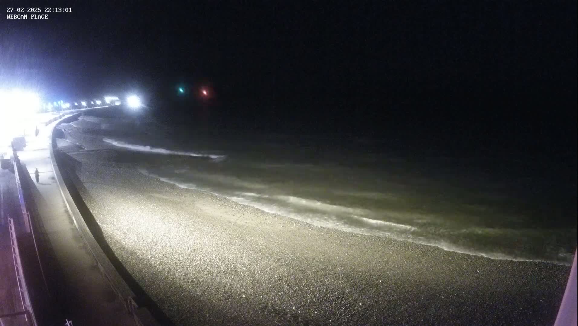 A dark, nighttime scene shows a pebble beach with gentle waves lapping the shore, under a cloudy sky.