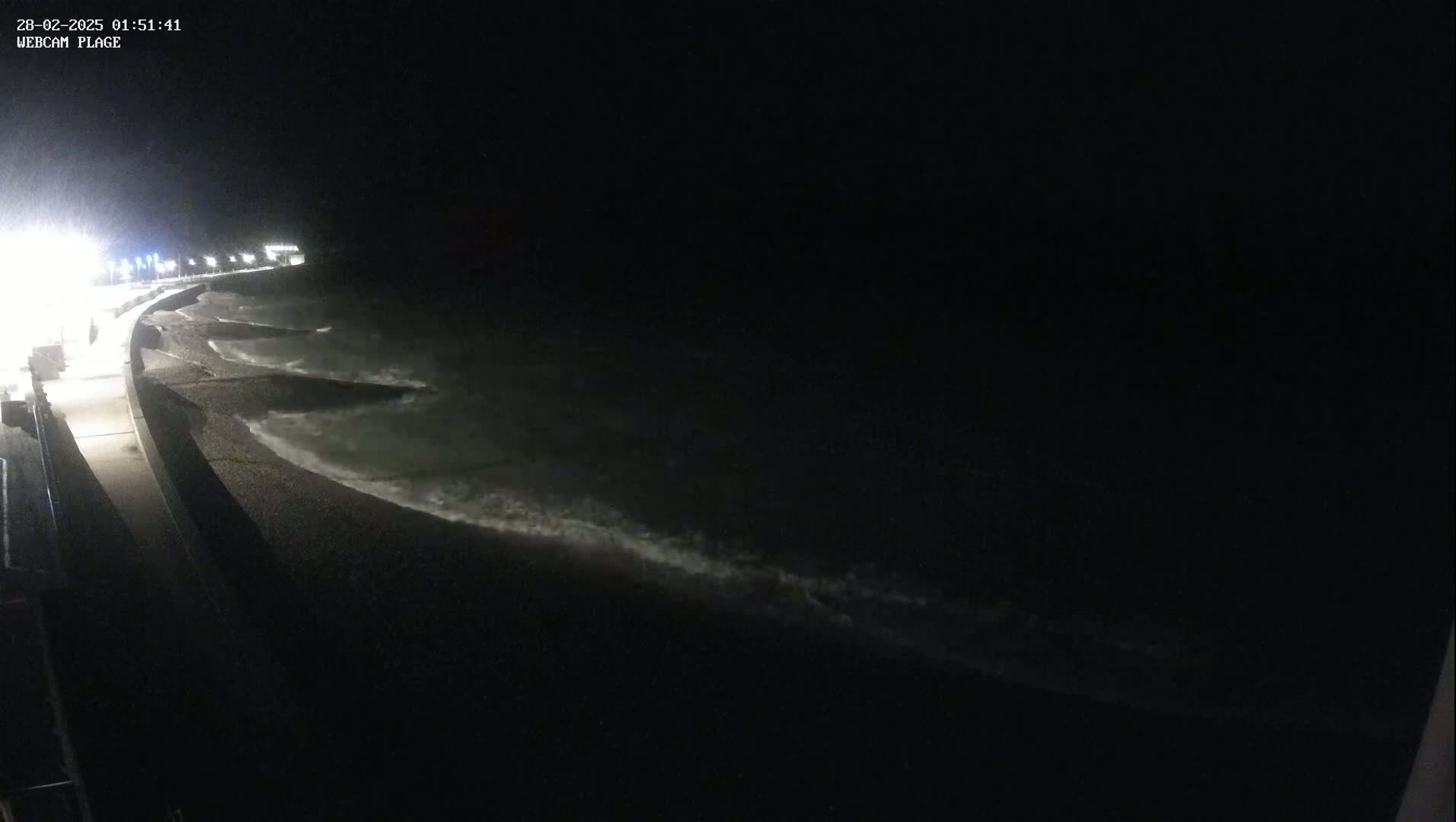Small waves lap a dark, pebble beach at night, illuminated by distant lights along a paved walkway.