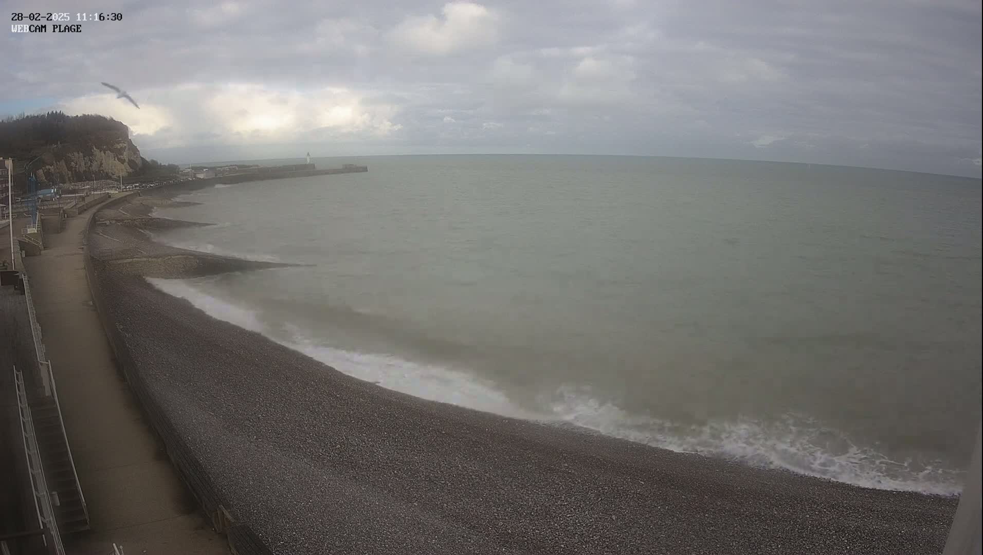 A pebble beach meets a calm, gray sea under a cloudy sky, with a cliff and a distant pier visible in the background.