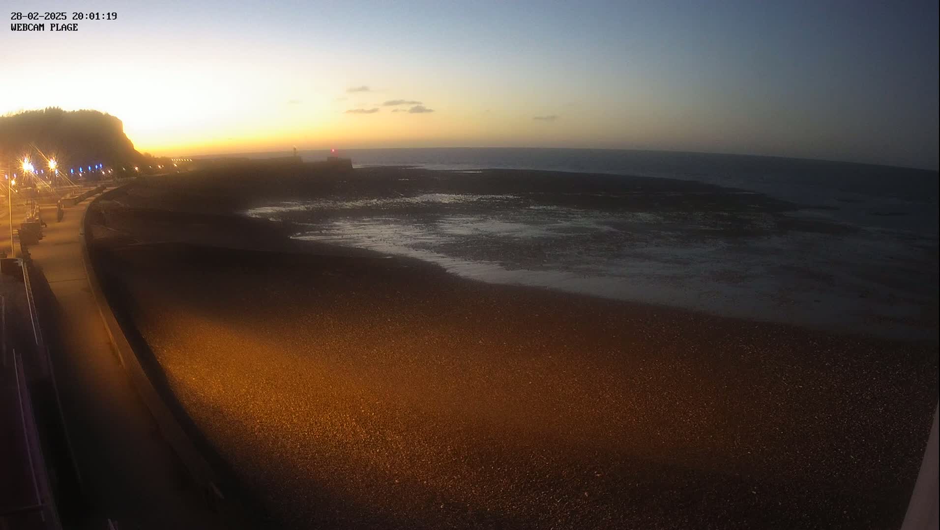 A dark, pebble beach is illuminated by artificial light at twilight, with a calm ocean and a distant cliff in the background.