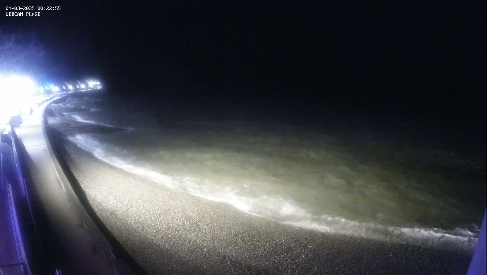 At night, small waves lap a pebbled shoreline next to a concrete wall under dim lighting.
