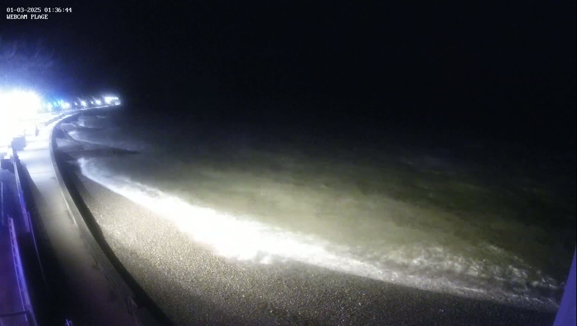 At night, small waves lap a pebble beach next to a seawall under artificial lighting.