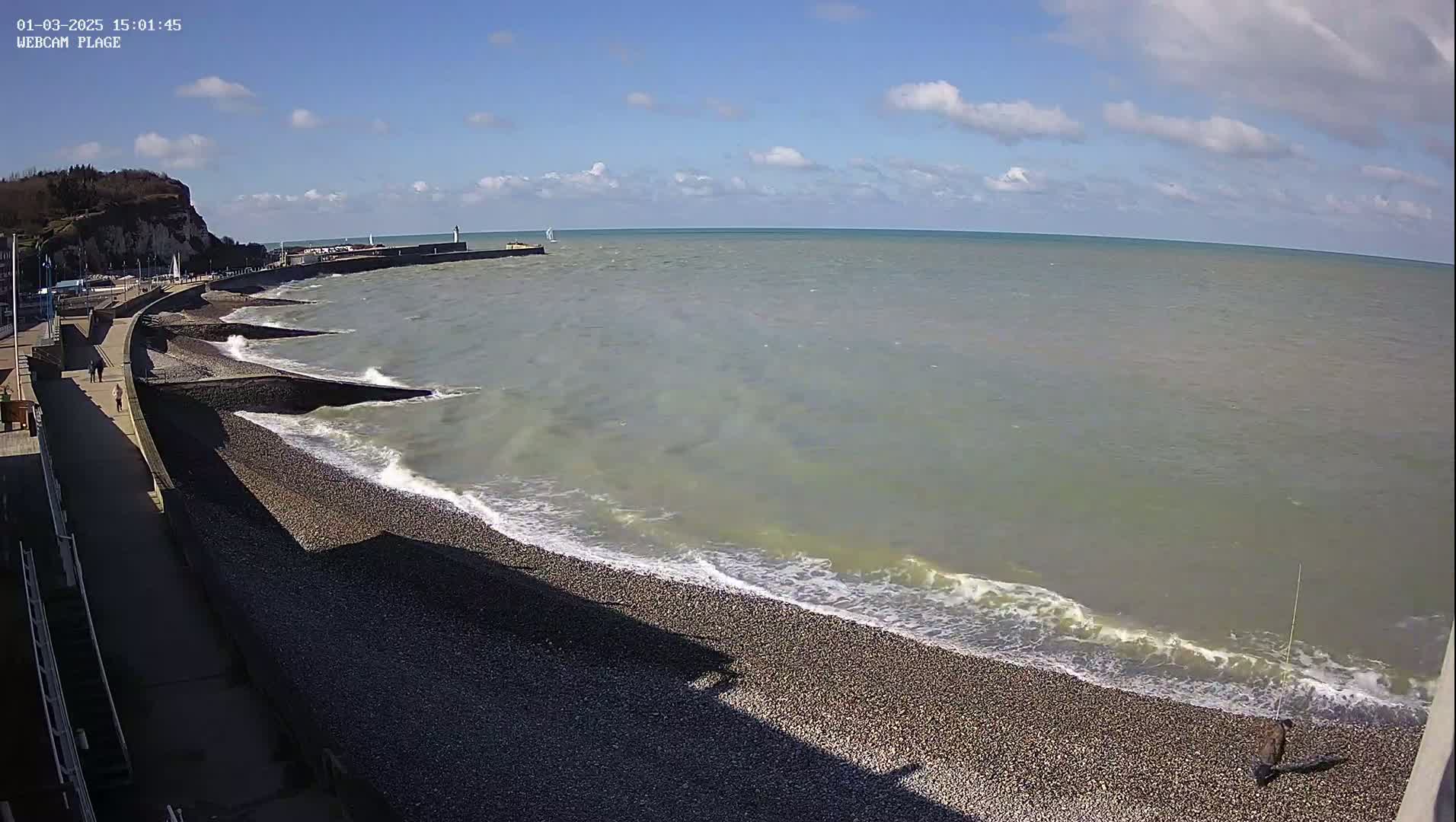 A rocky beach with moderate waves under a partly cloudy sky stretches out from a paved walkway and cliffside.