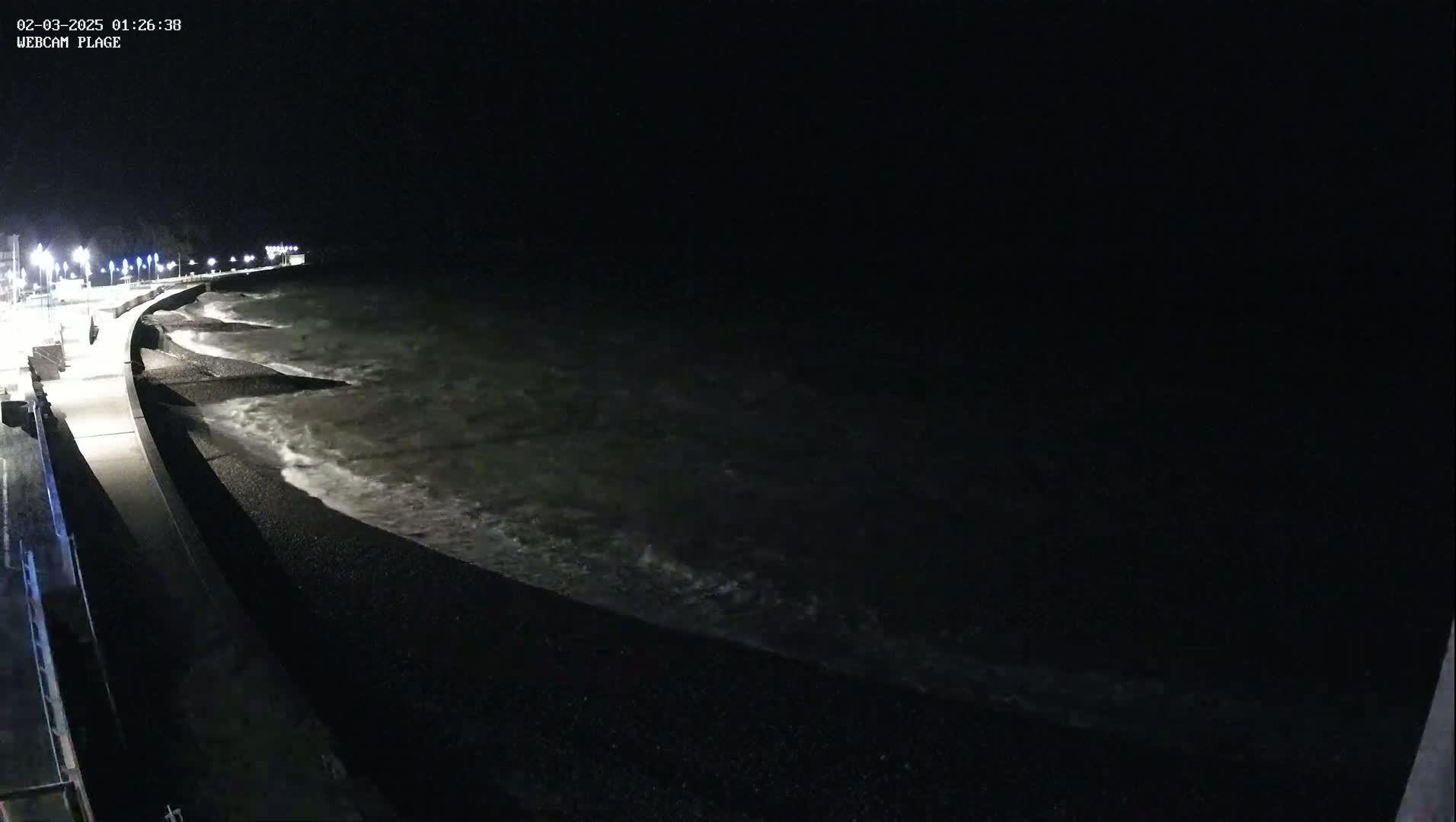 A dark, nighttime view of a pebble beach with waves lapping the shore and a paved walkway lined with lights extending into the distance.