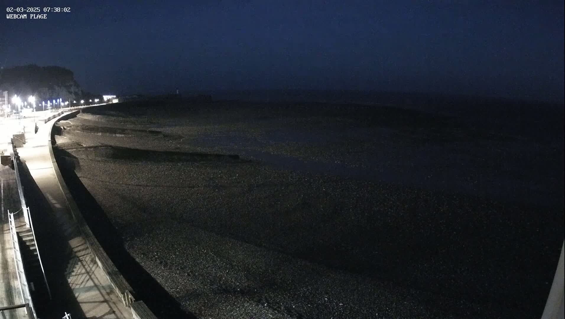 A dark, nighttime view of a pebble beach and a paved walkway illuminated by streetlights, under a mostly clear, dark sky.
