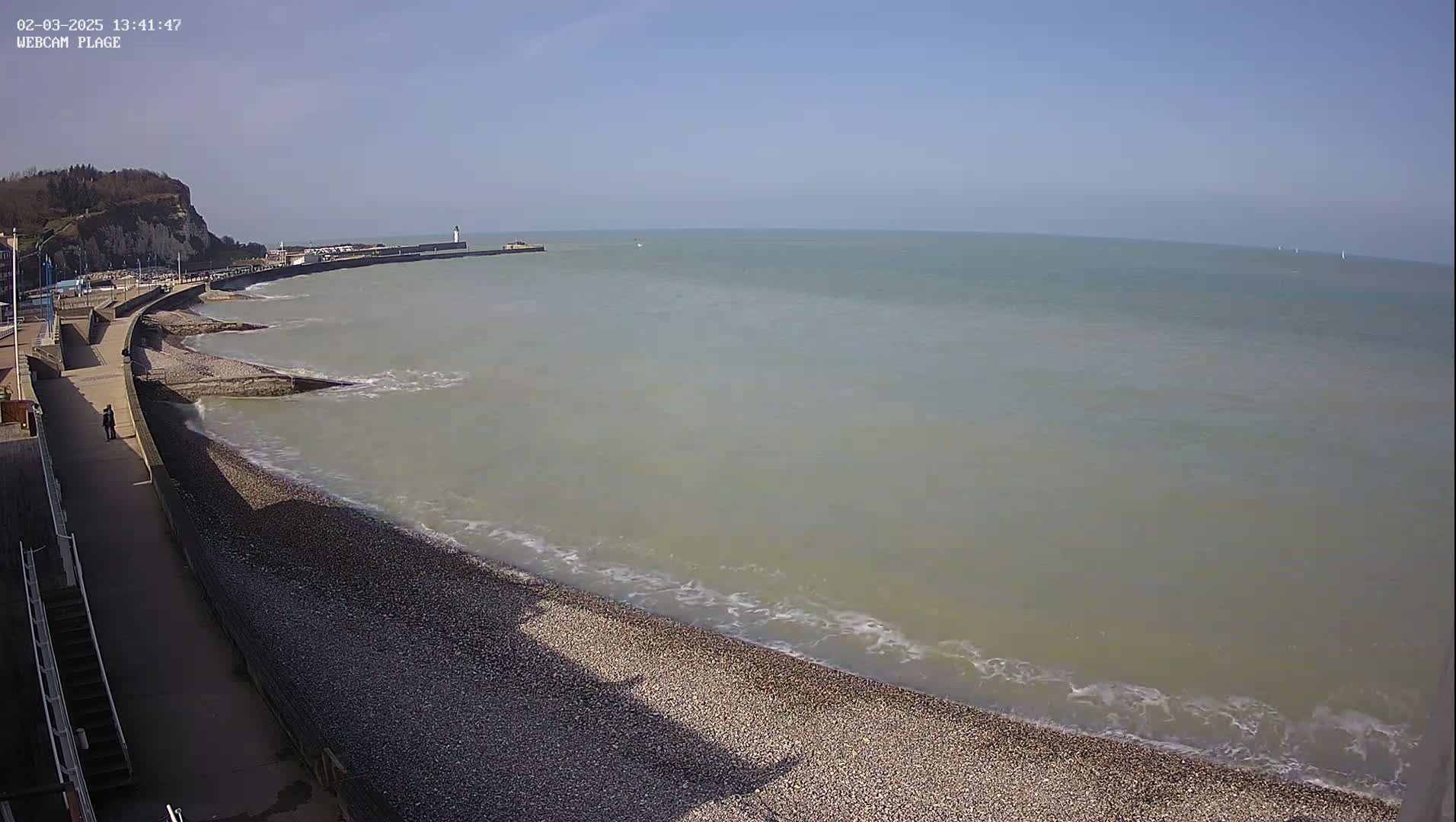 A pebble beach curves alongside a walkway, meeting a calm, light-grey sea under a clear blue sky.