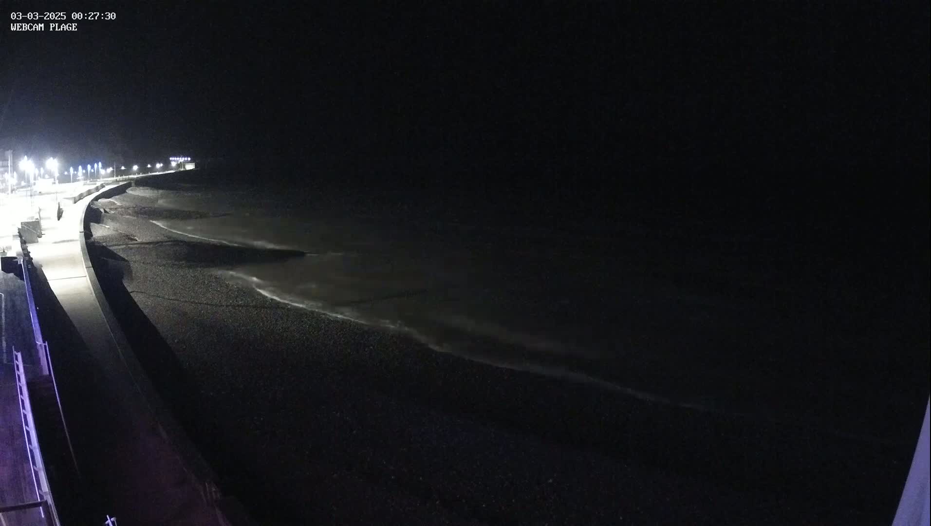 A dark, nighttime view shows a pebble beach and a paved walkway lit by streetlights, with gentle waves lapping the shore.
