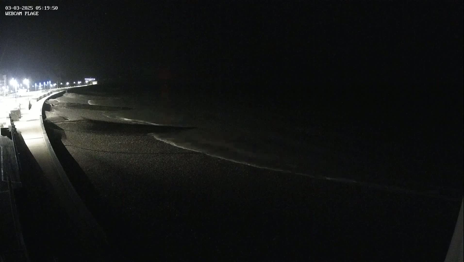 A dark, nighttime beach scene shows gently lapping waves on a dark-colored, pebbly shore next to a lit walkway.