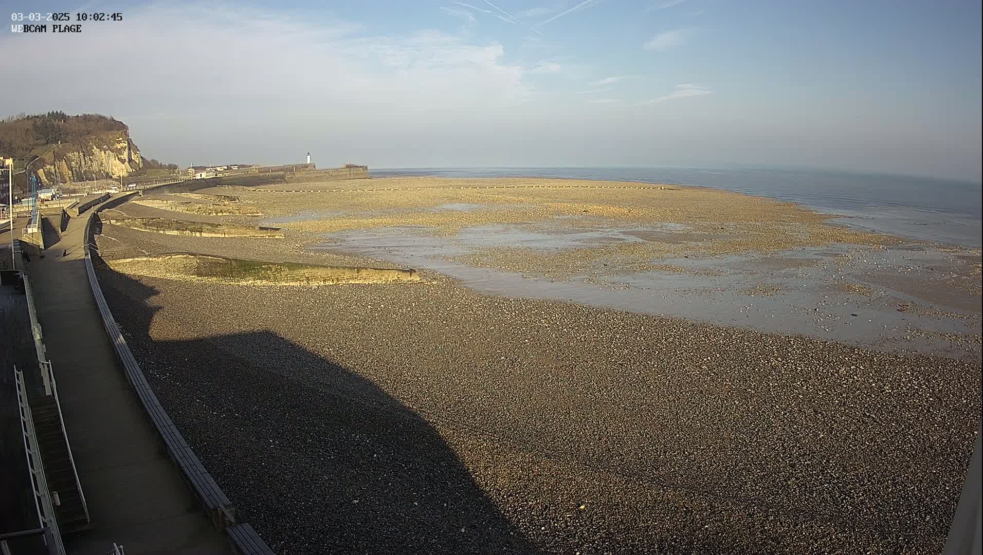 A mostly sunny day reveals a pebbled beach at low tide, with a paved walkway and a cliff in the background.
