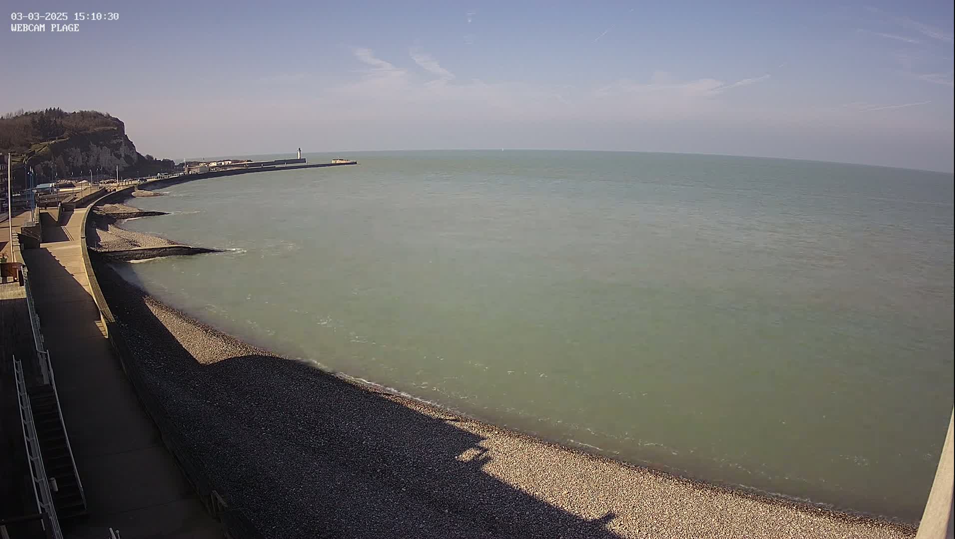 A pebble beach curves along a calm, greenish-grey sea under a clear blue sky.