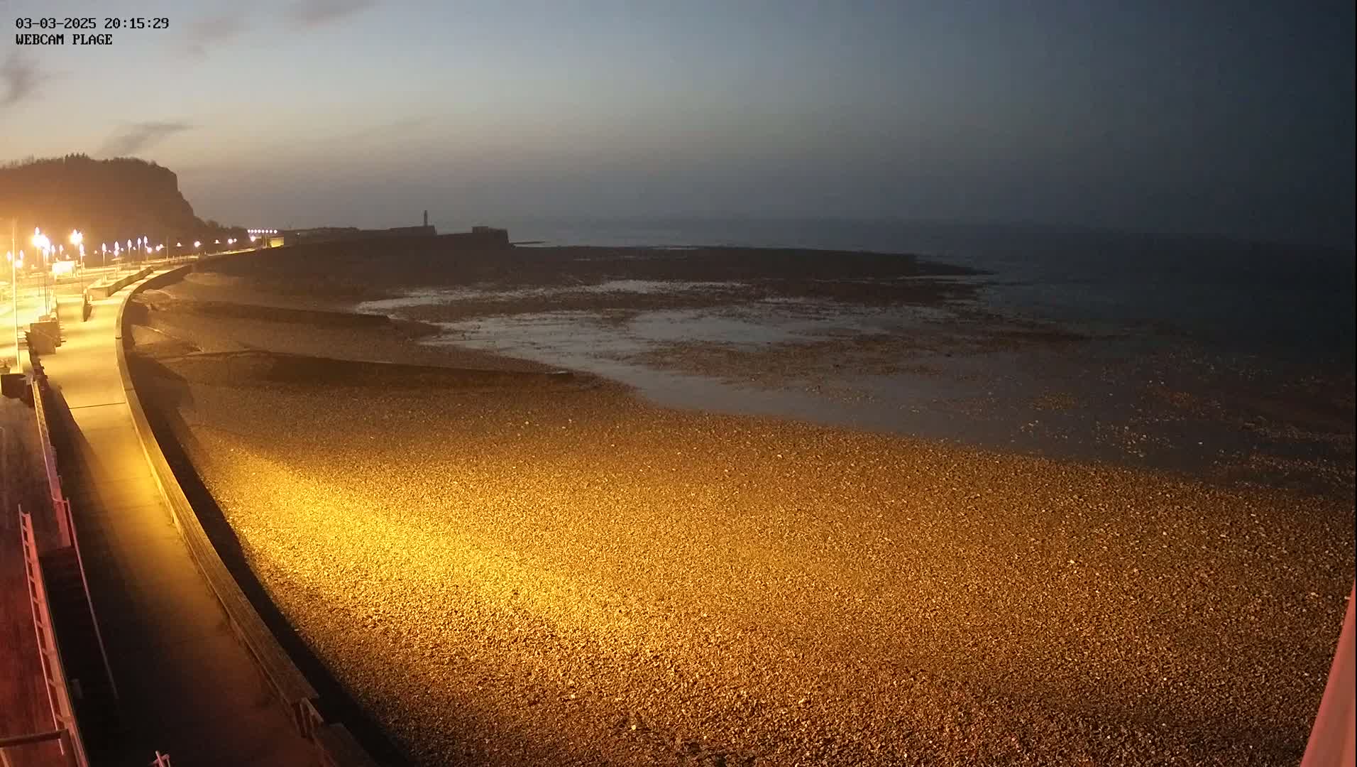 A dark, pebble beach is illuminated by artificial lights at night, with a calm sea and a distant hill visible in the background.