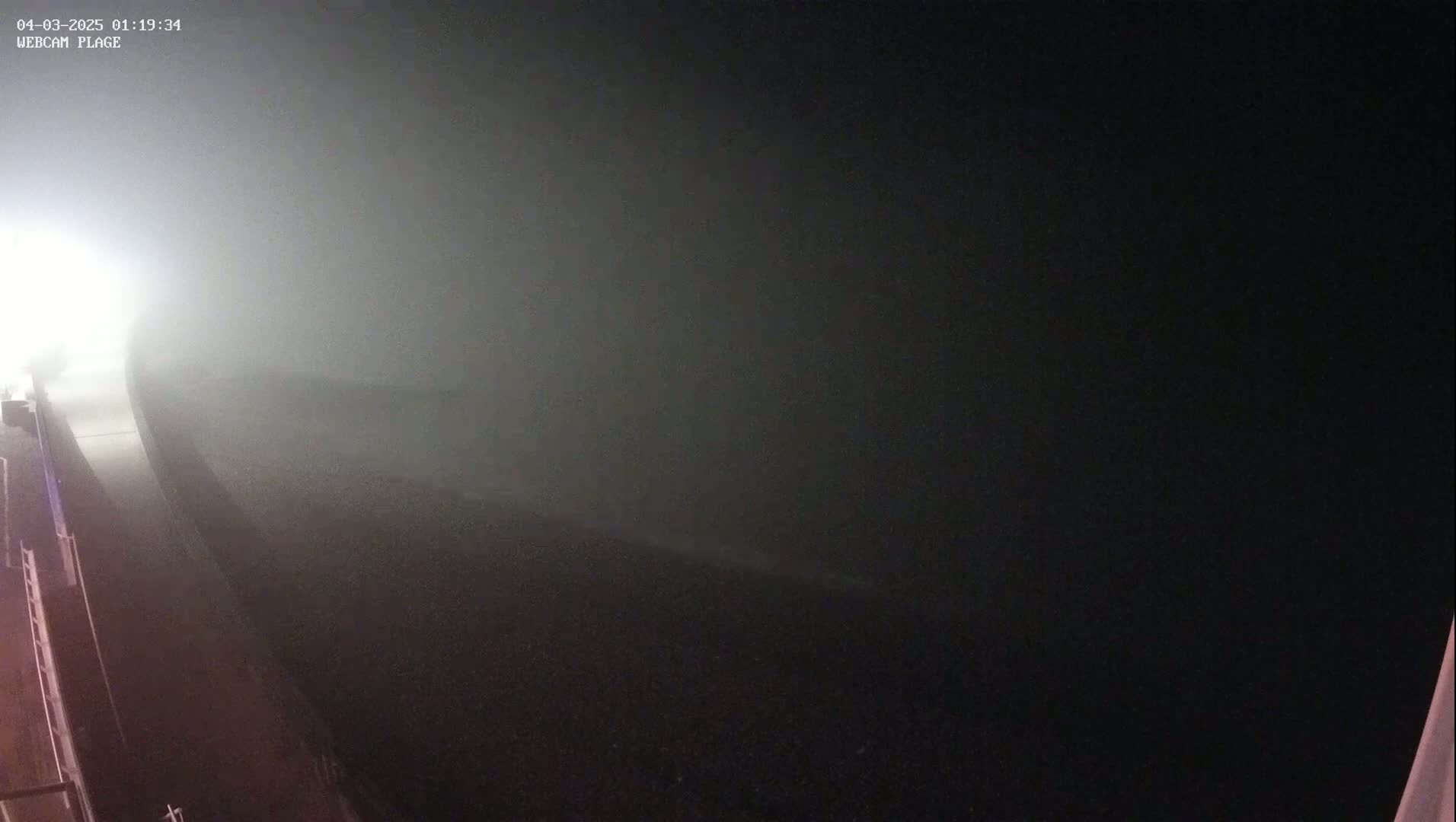 A dark road is shrouded in thick fog at night, illuminated only by a bright light source in the distance.