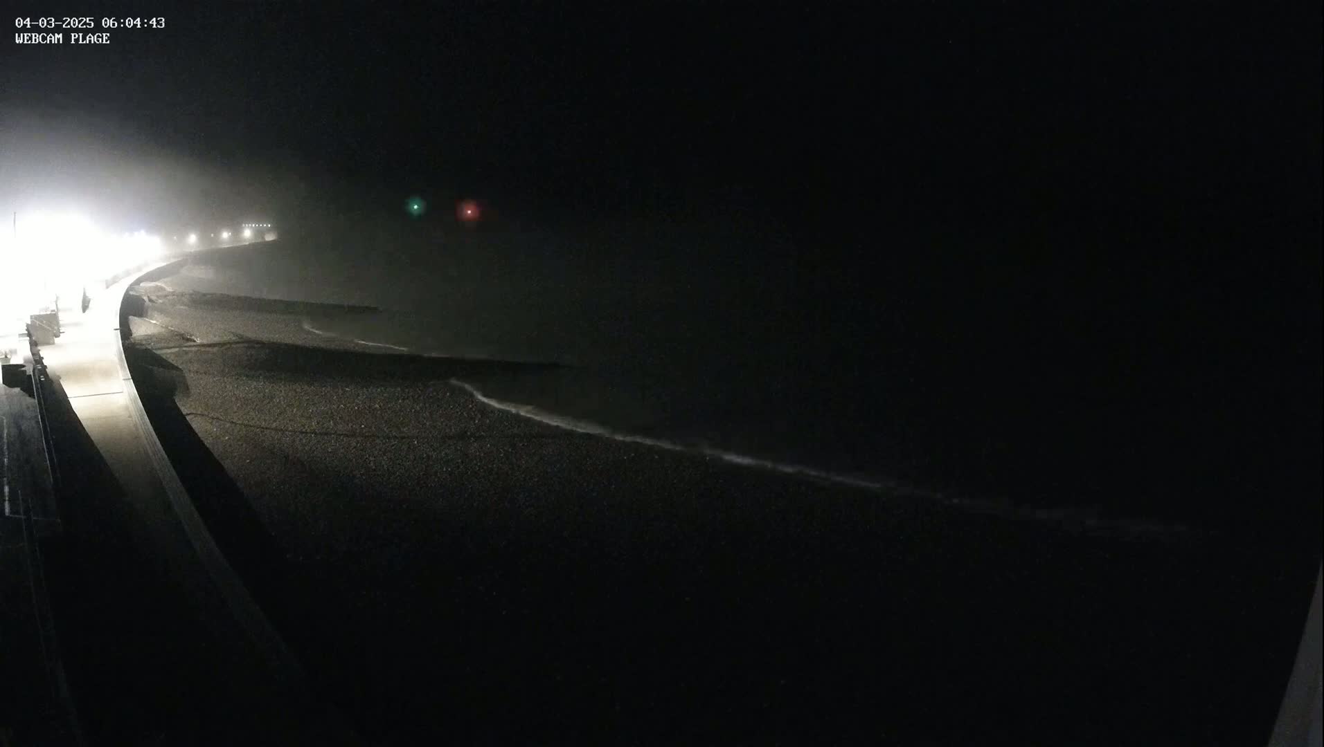 A dark, foggy night scene shows a shoreline with gentle waves lapping at a pebble beach, illuminated by distant lights.