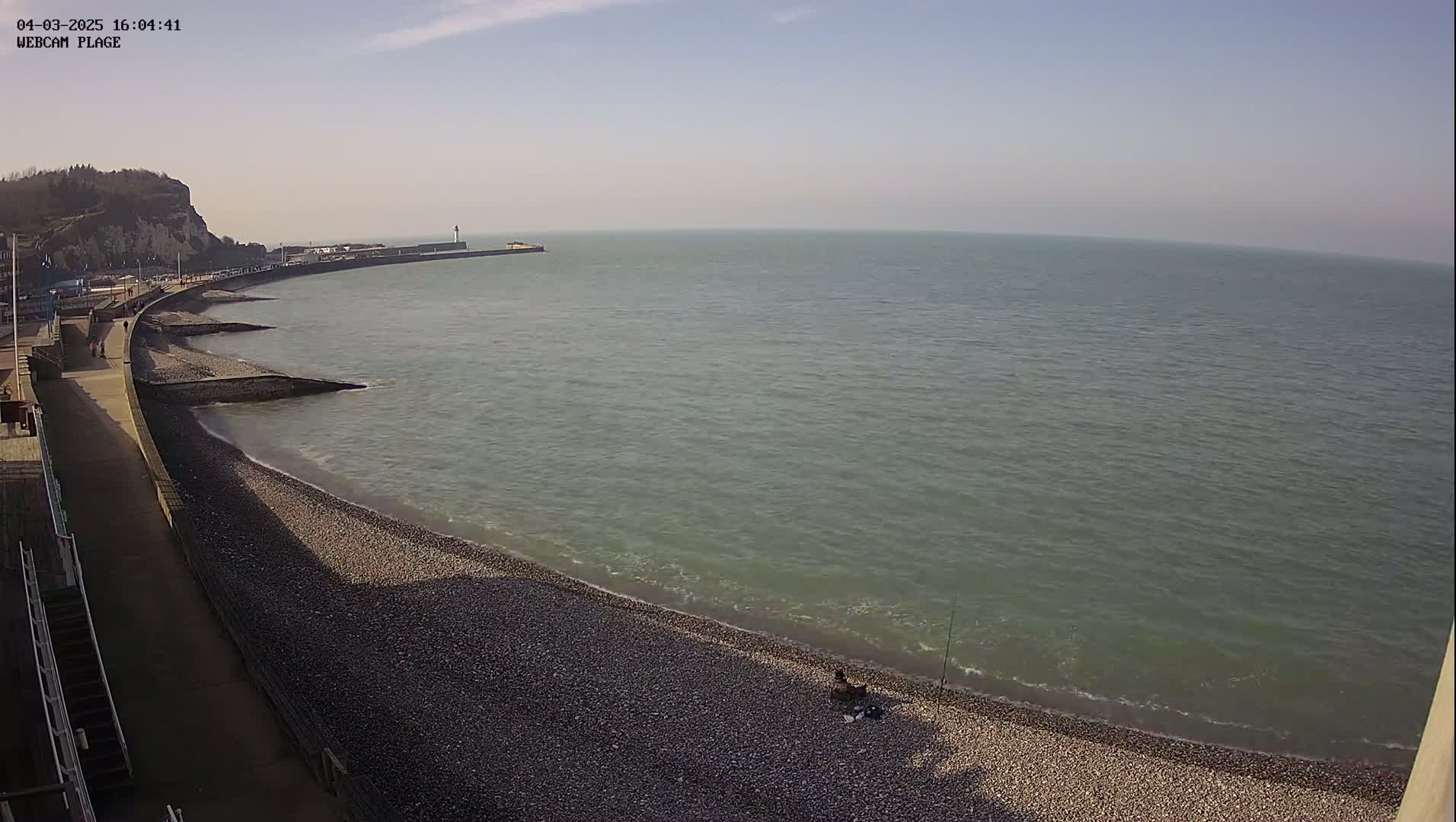 A pebble beach curves along a calm sea under a mostly clear sky, with a walkway and distant cliffs visible.
