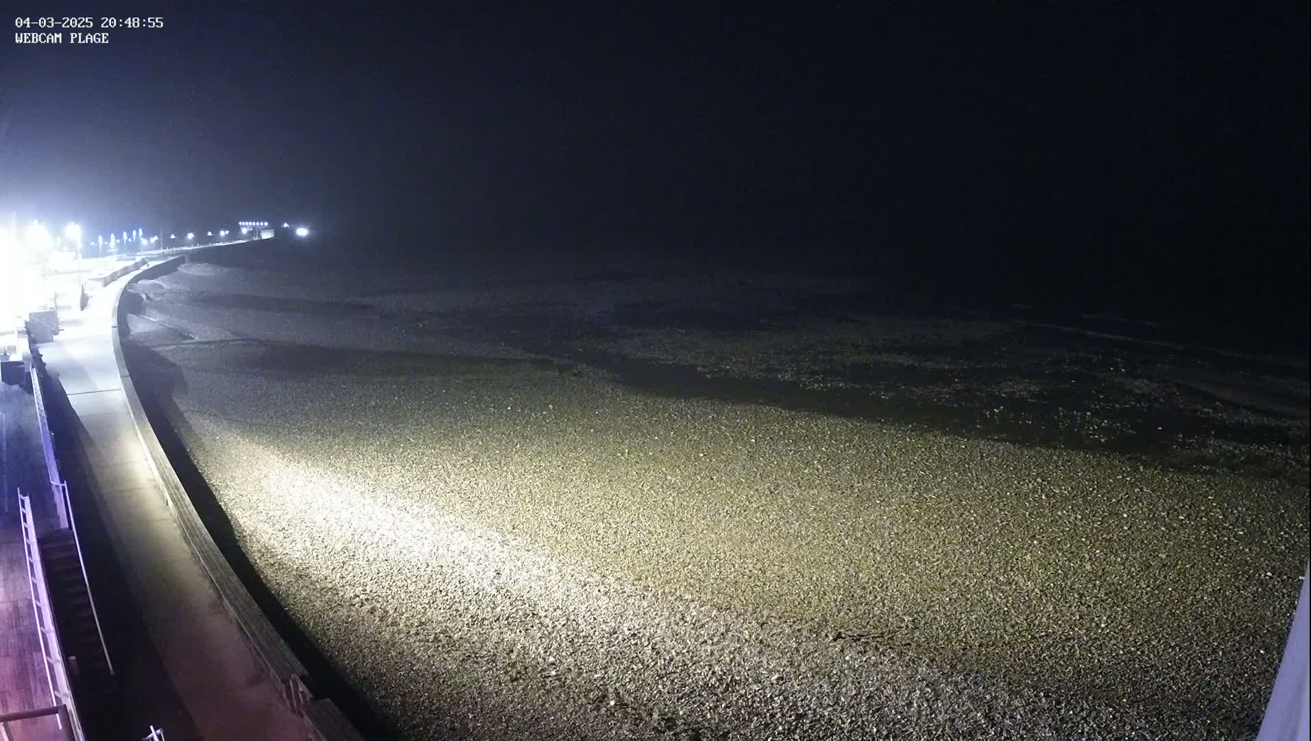 A nighttime view of a pebble beach and a paved walkway, illuminated by artificial lights, under a dark sky.