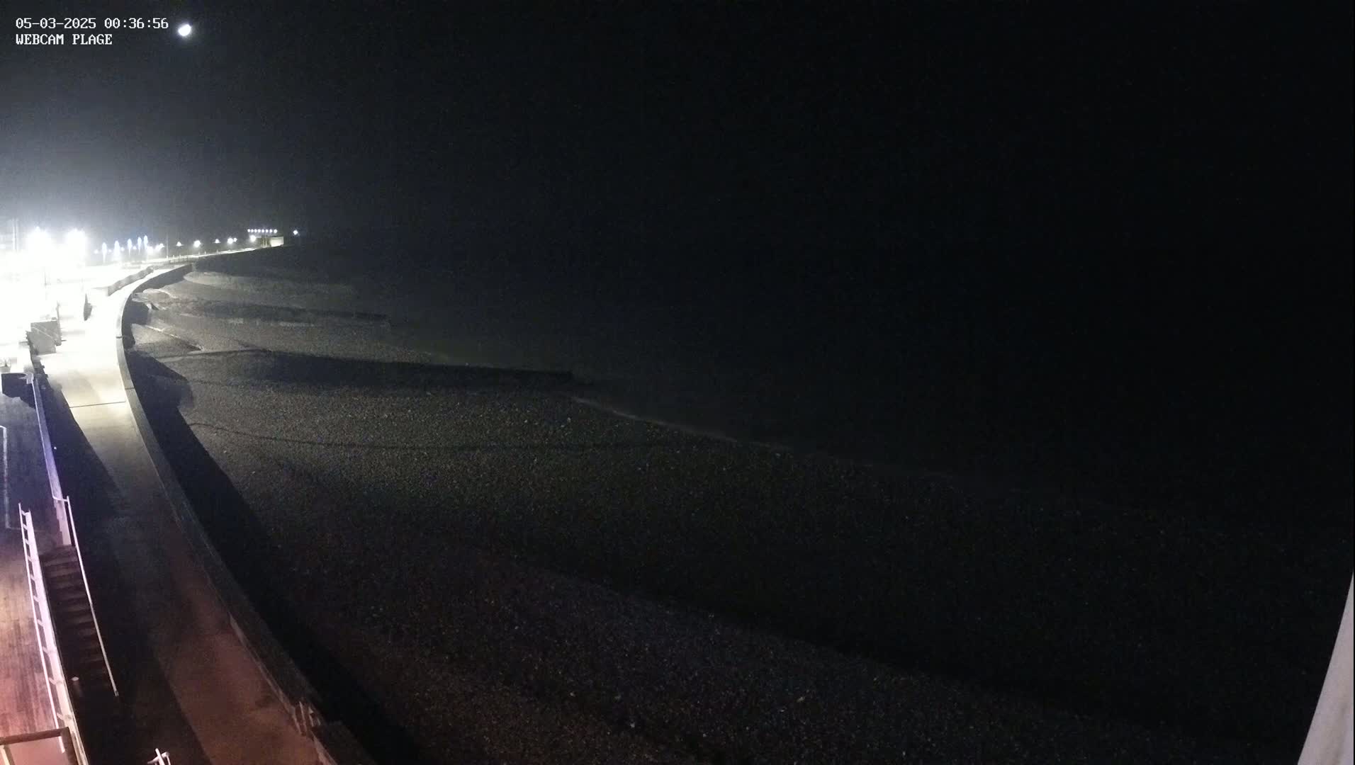 A dark, nighttime view of a pebble beach and a paved walkway, illuminated by streetlights, with gentle waves visible near the shore under a clear sky.