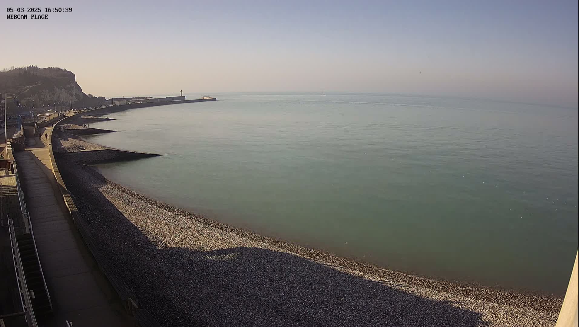 A pebble beach curves along a sea wall under a clear, bright sky, with calm, greenish-grey water extending to the horizon.
