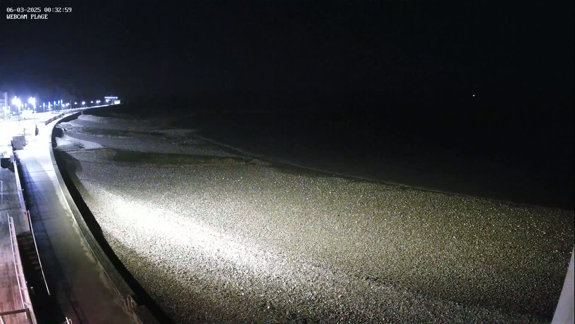 A dark, nighttime view of a pebble beach next to a paved walkway, illuminated by artificial lights.