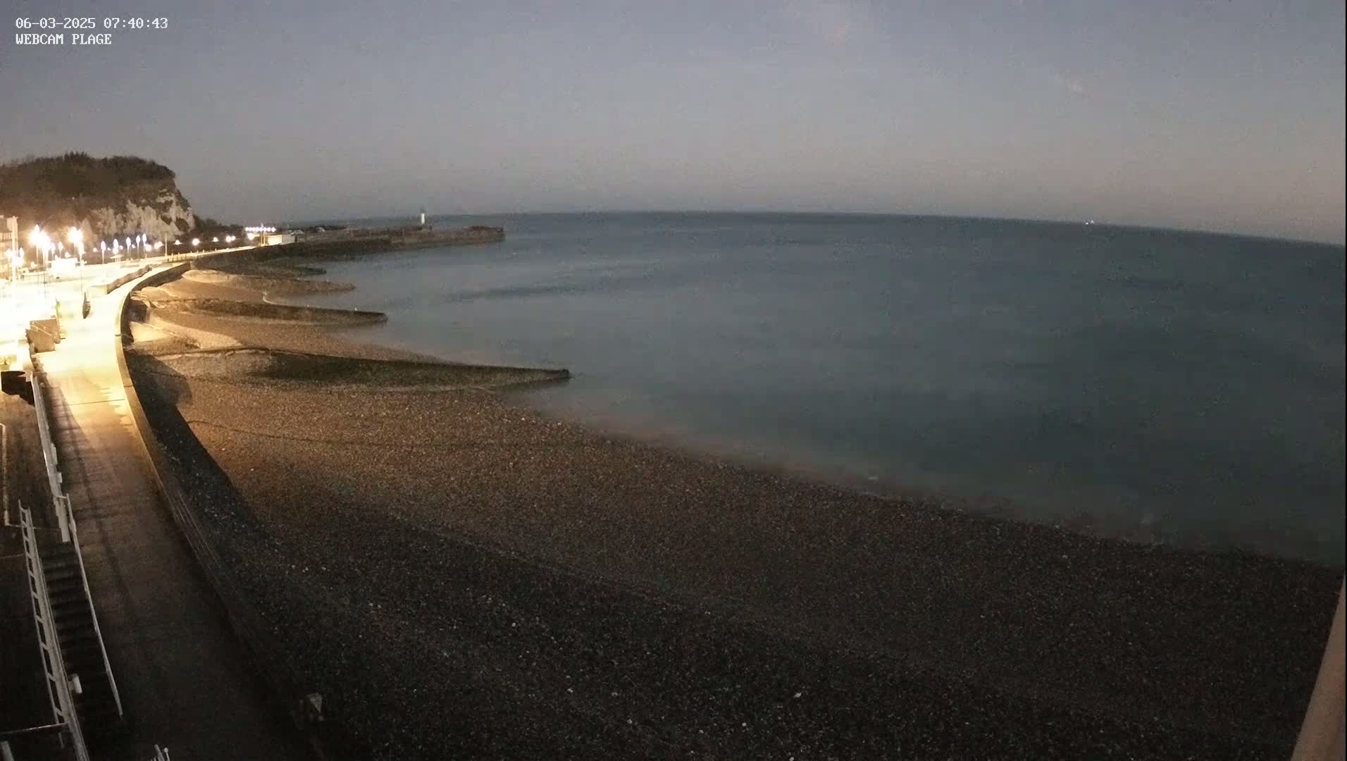 A dark, pebble beach and a paved walkway are illuminated at night next to a calm, dark-grey sea under a dusky sky.