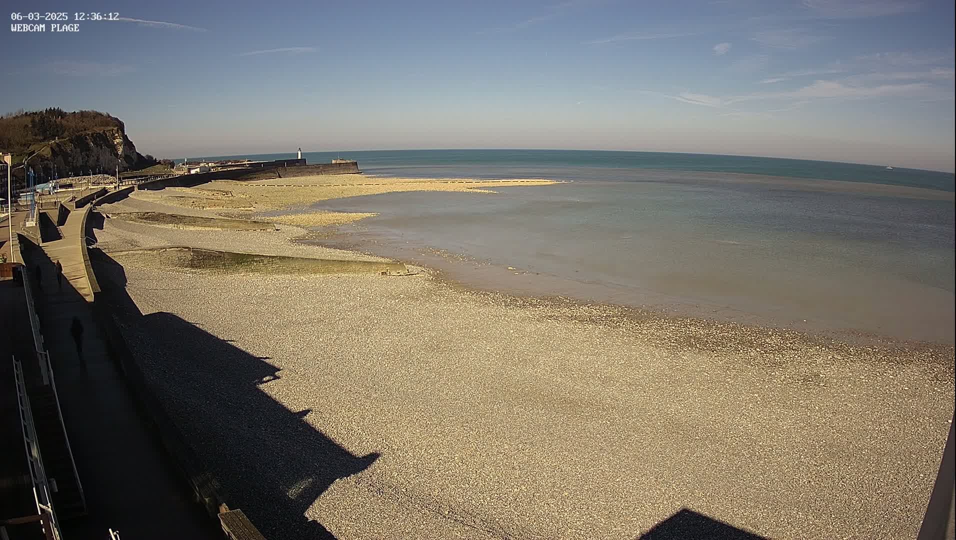 A pebble beach stretches to a calm, light-brown sea under a clear blue sky.