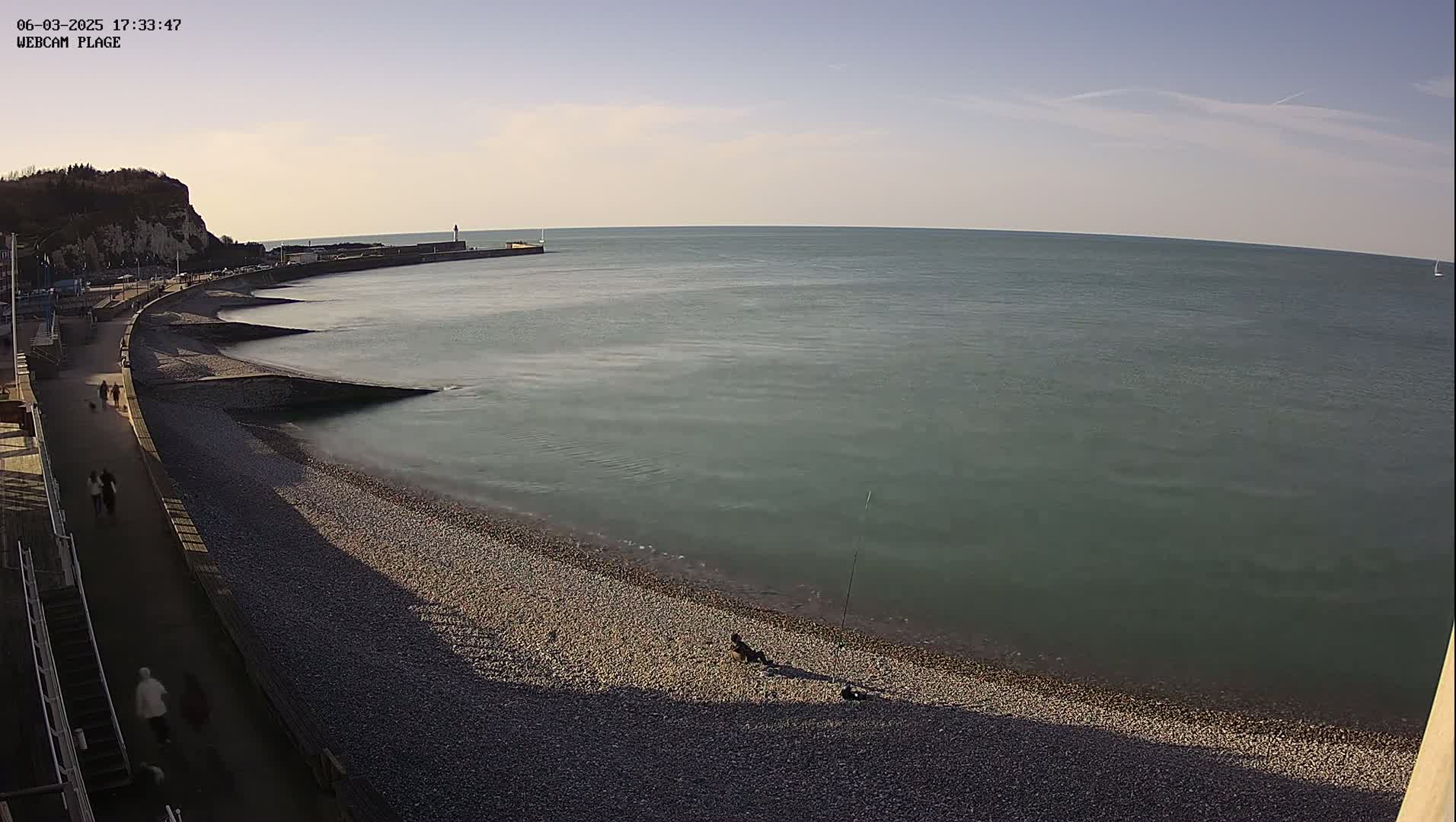 A pebble beach curves along a calm, teal sea under a mostly clear sky, with a few people walking along a paved path and one person fishing near the water's edge.