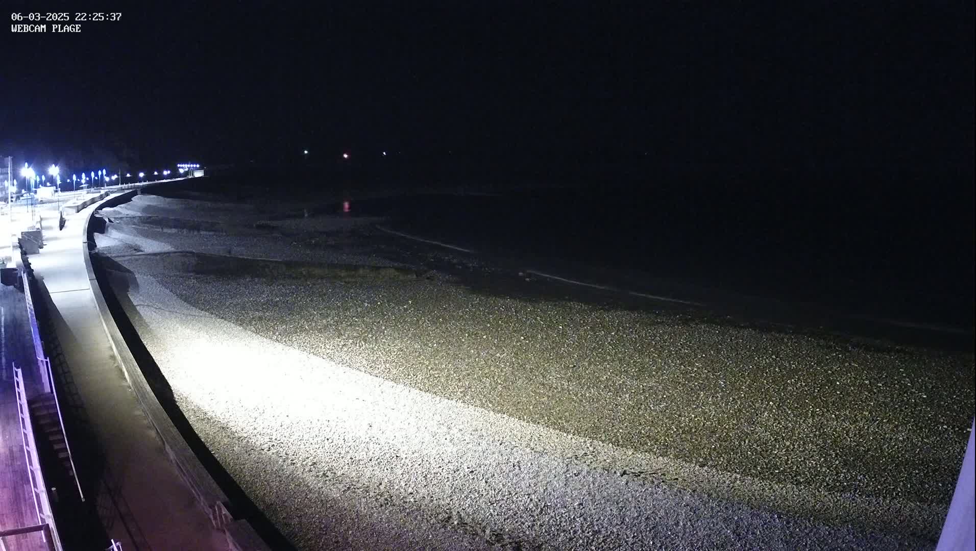 A paved walkway curves along a dark, pebble beach at night next to a calm, dark body of water.