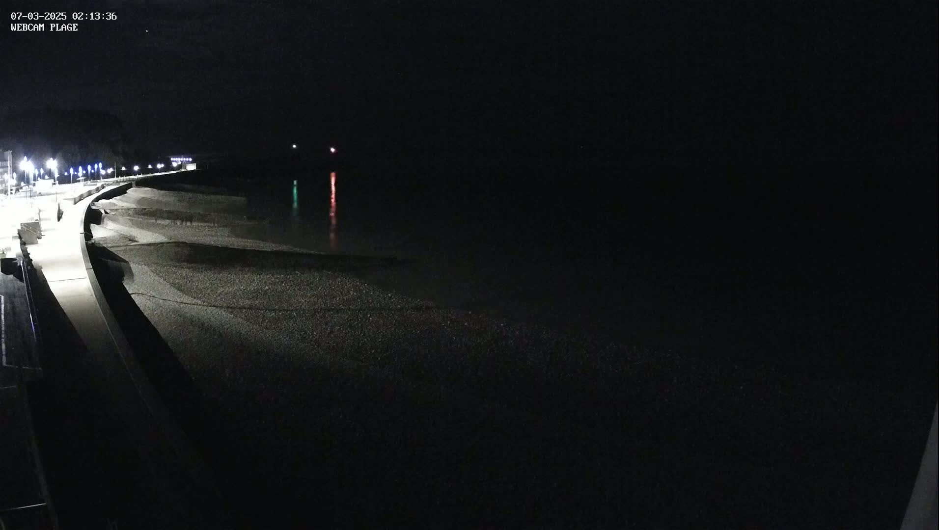 A dark, nighttime view of a pebble beach and a paved walkway illuminated by streetlights, with calm water and faintly visible lights in the distance.
