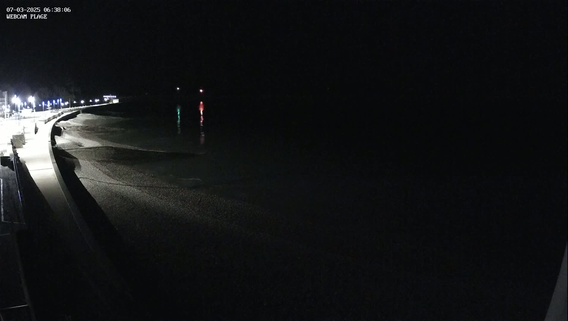 A dark, nighttime view shows a paved walkway along a pebble beach with gently lapping waves and faintly visible lights in the distance.