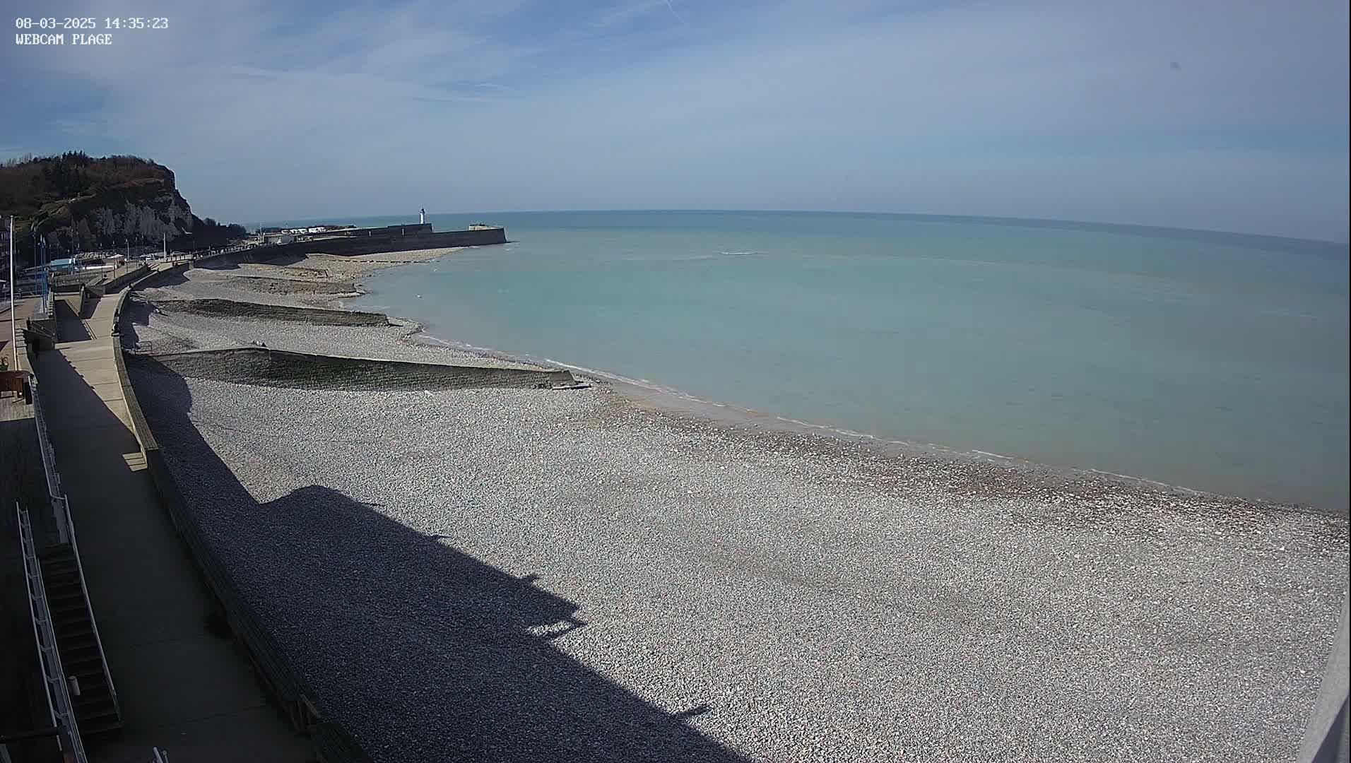 A pebble beach stretches along a calm, light-blue ocean under a clear, sunny sky, with a walkway and cliff visible in the background.